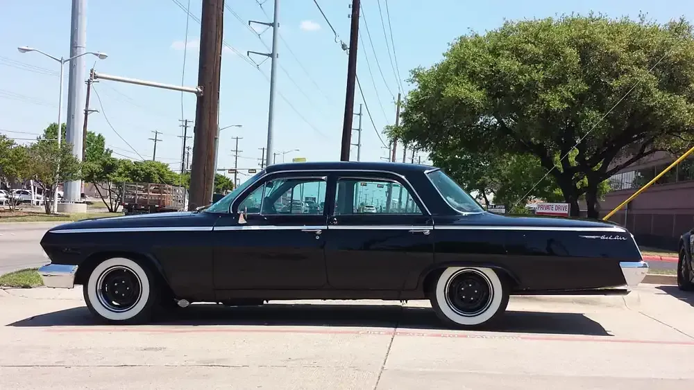 A side view of a black classic Chevrolet sedan with white-wall tires parked on pavement under a sunny, blue sky.
