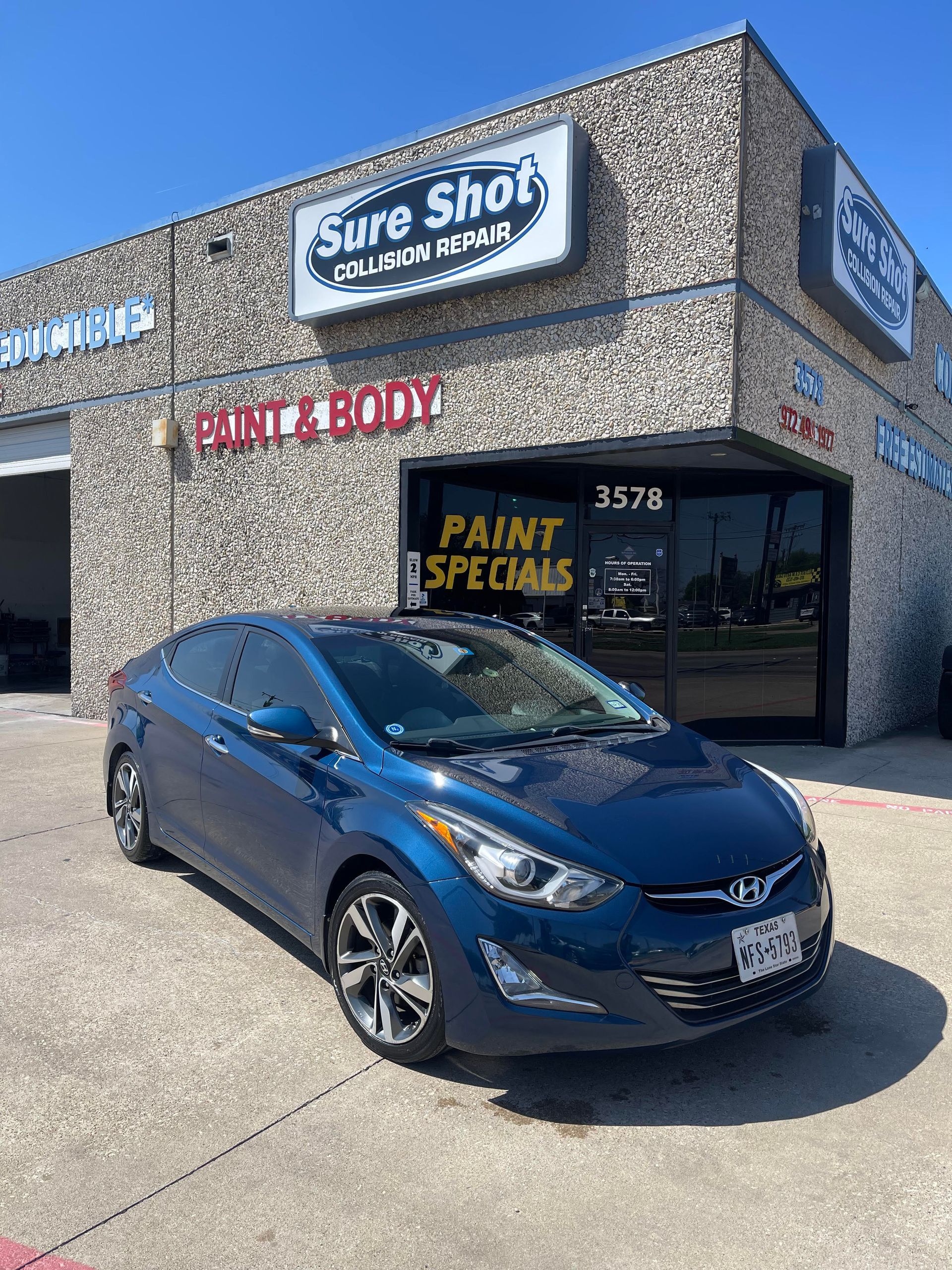A blue Hyundai sedan parked in front of a Sure Shot Paint & Body auto repair shop on a sunny day.