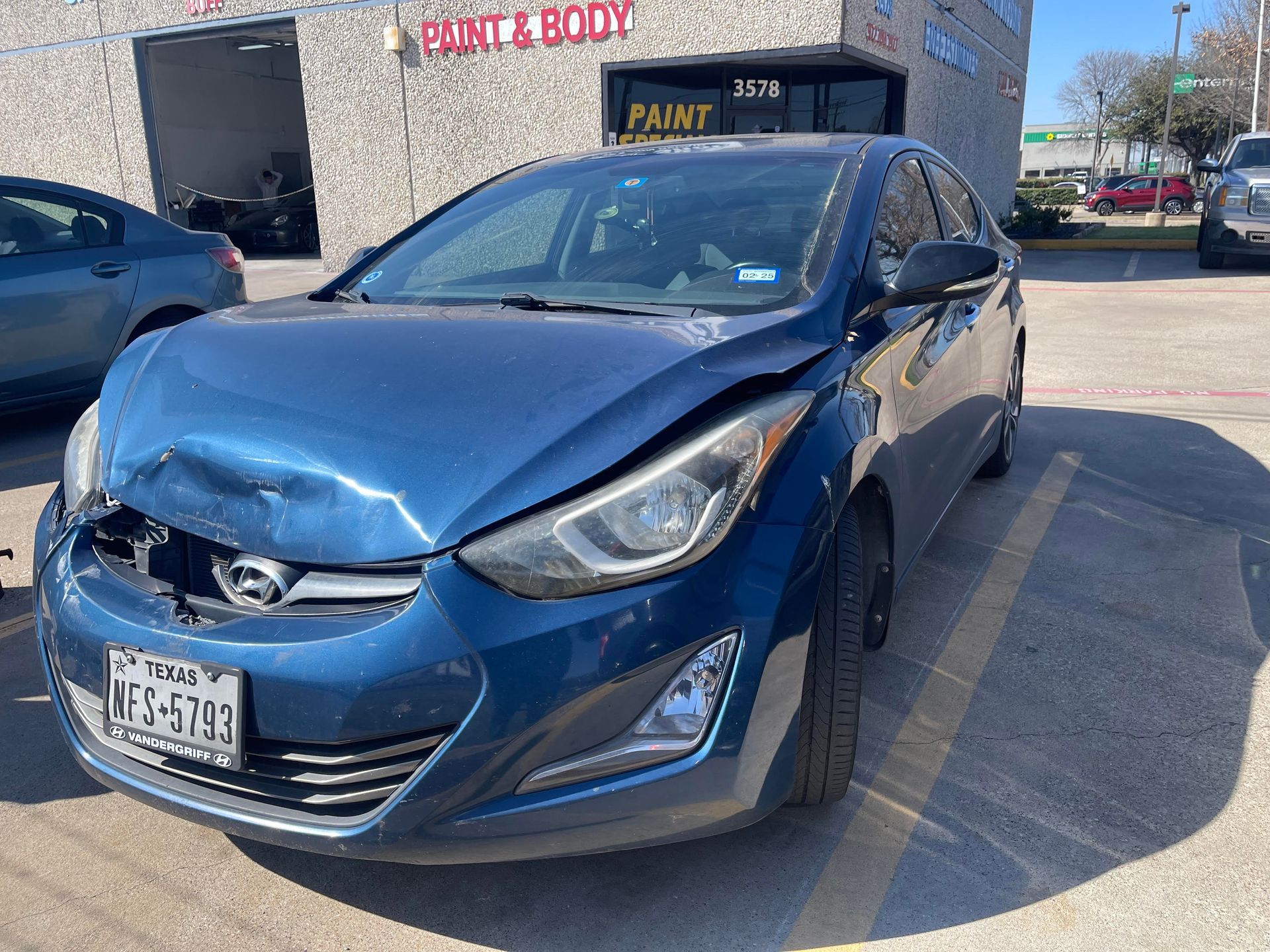 A blue Hyundai sedan with significant front-end collision damage parked in a lot in front of an auto body shop.
