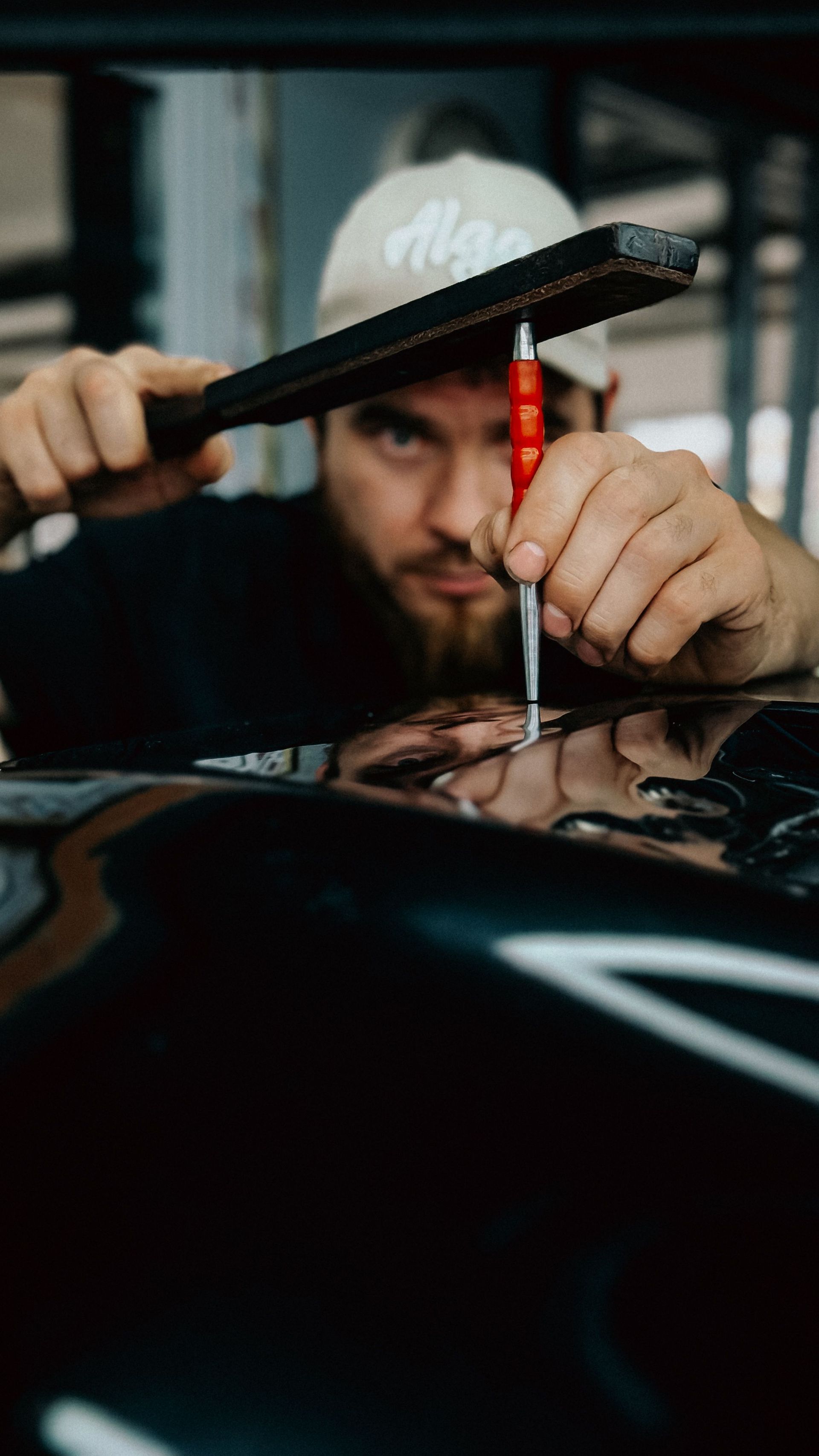 Mechanic inspecting a black car’s surface with a tool, seen through a windshield in a dim garage
