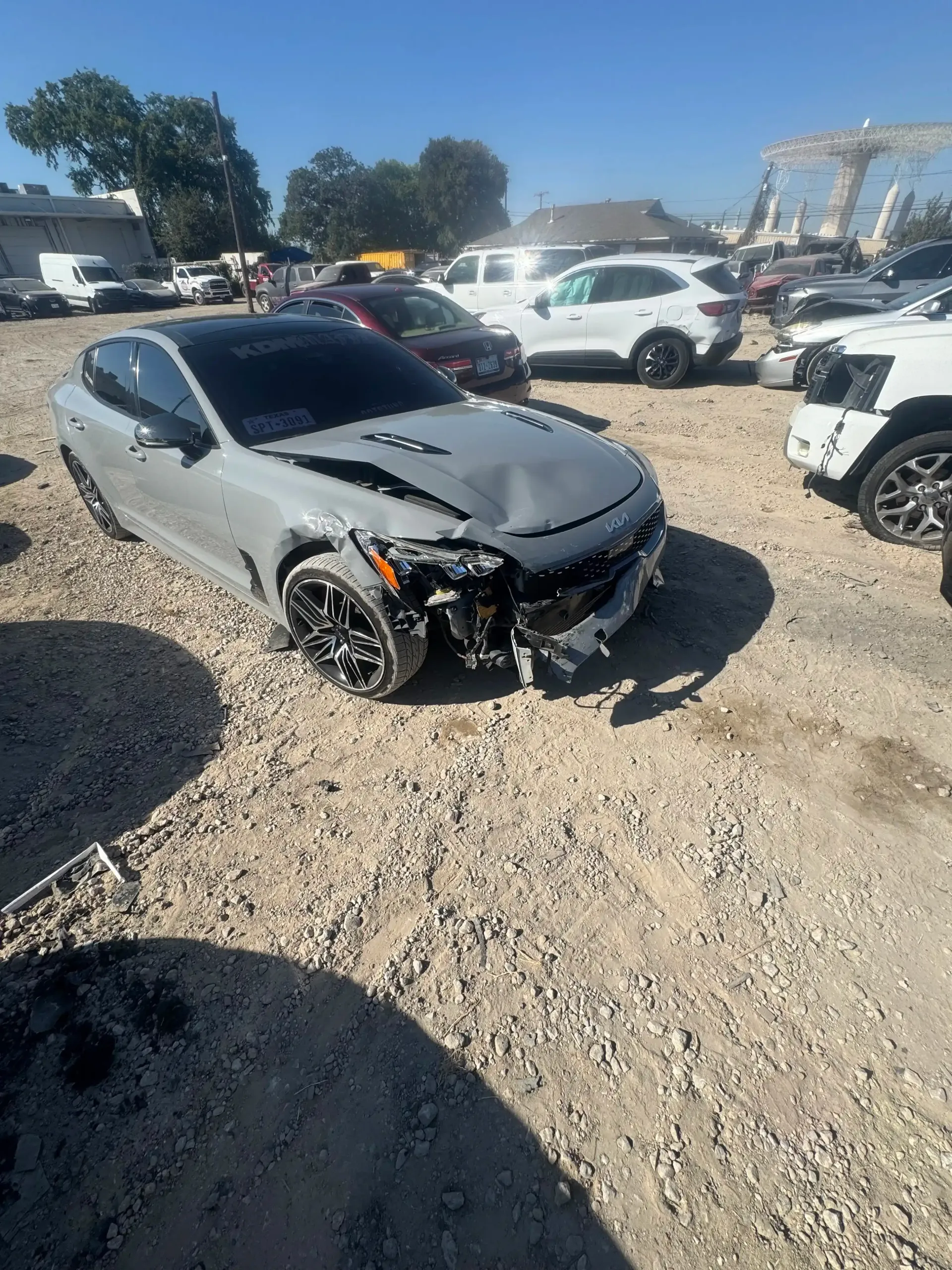 A damaged grey Kia Stinger parked in a gravel lot among other vehicles on a sunny day.