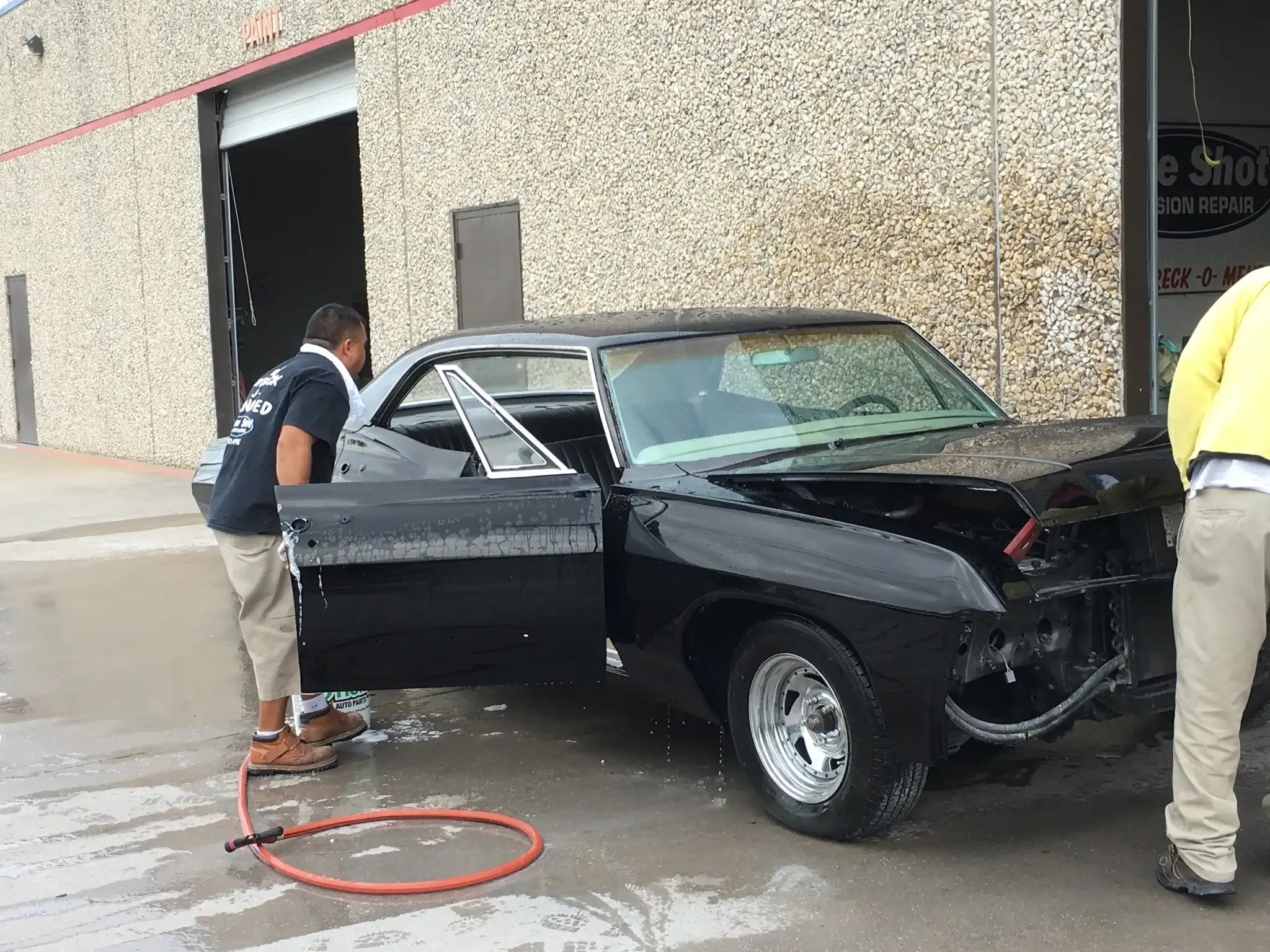 Two people wash a black vintage car parked outside a building with a textured, light-colored facade.