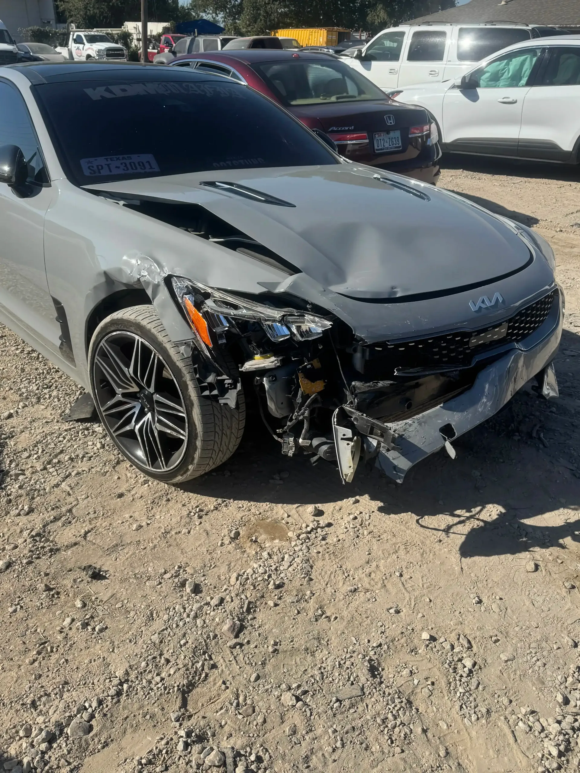 A damaged gray Kia Stinger with a smashed front bumper and hood parked on a dirt lot with other cars in the background.