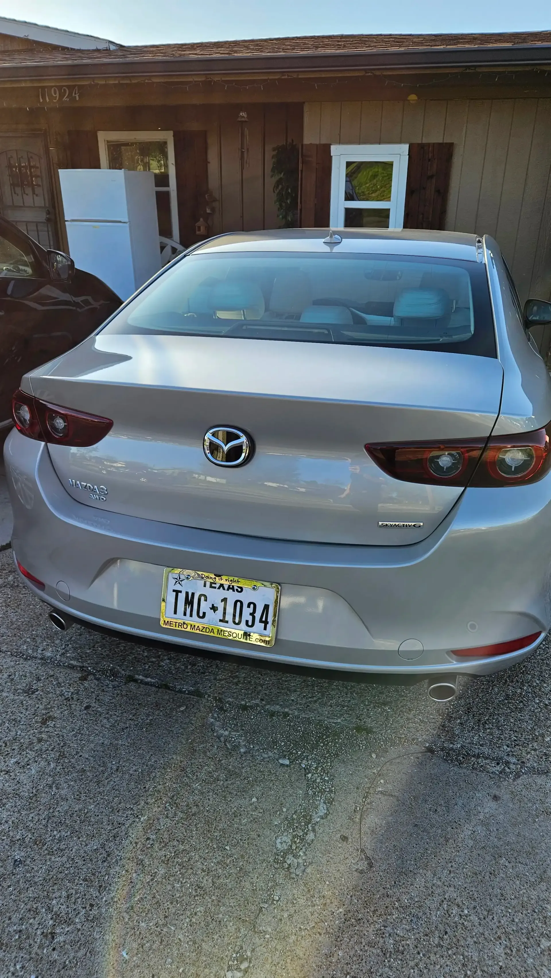A silver Toyota Corolla parked on a gravel driveway in front of a house with a white appliance visible in the background.