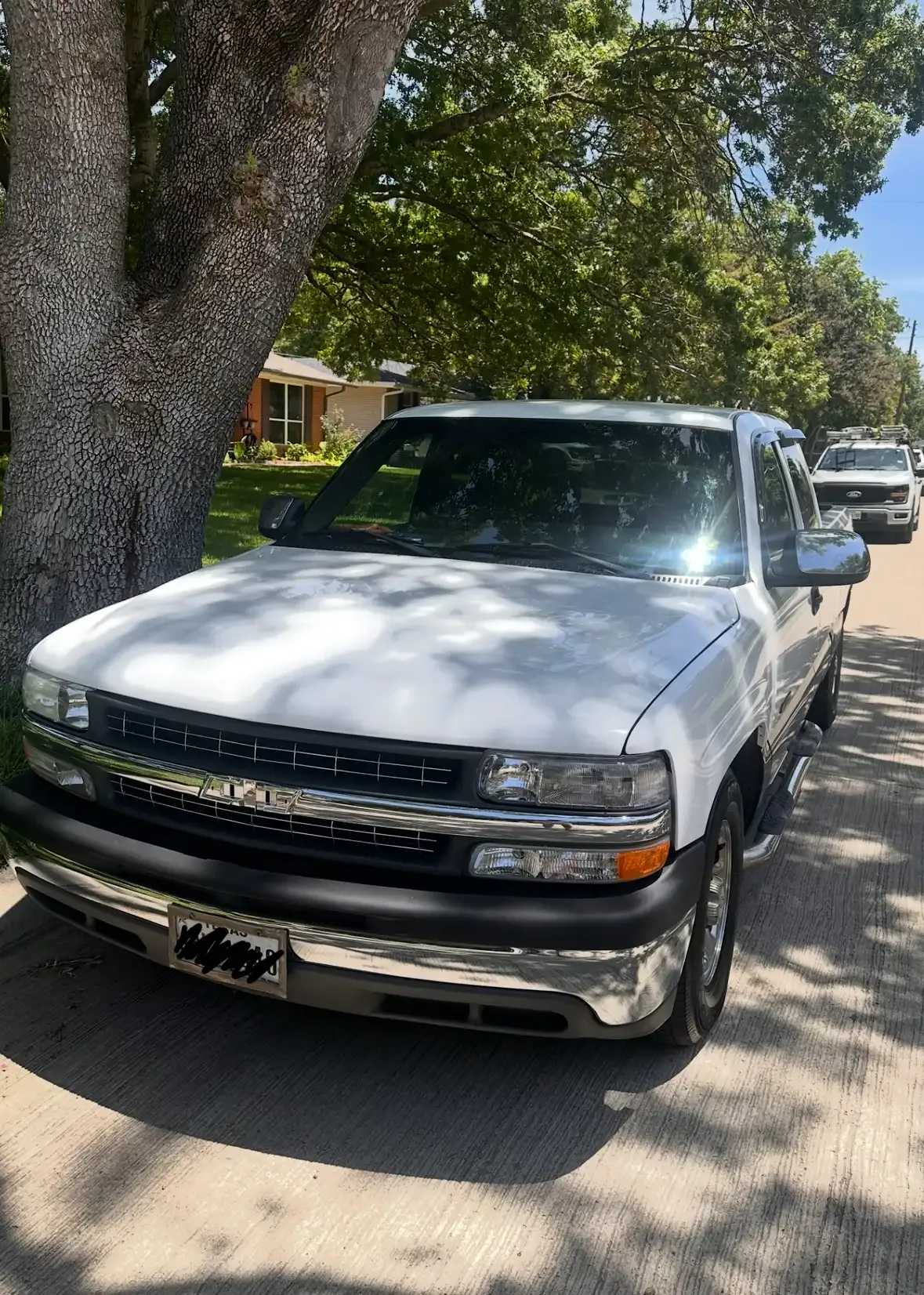 A white pickup truck parked on a residential street next to a large tree.