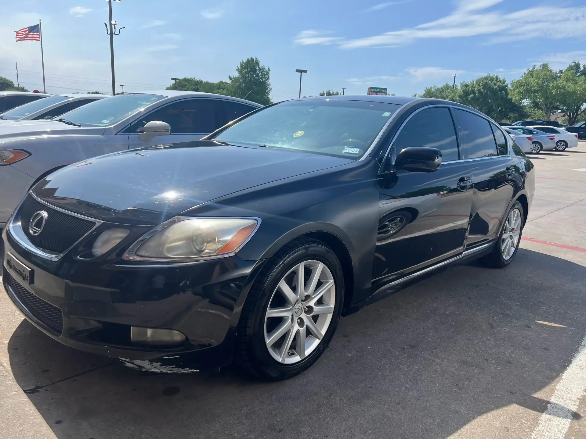 A black Lexus sedan parked in a sunny outdoor lot, angled toward the front, with a partially visible car behind it.