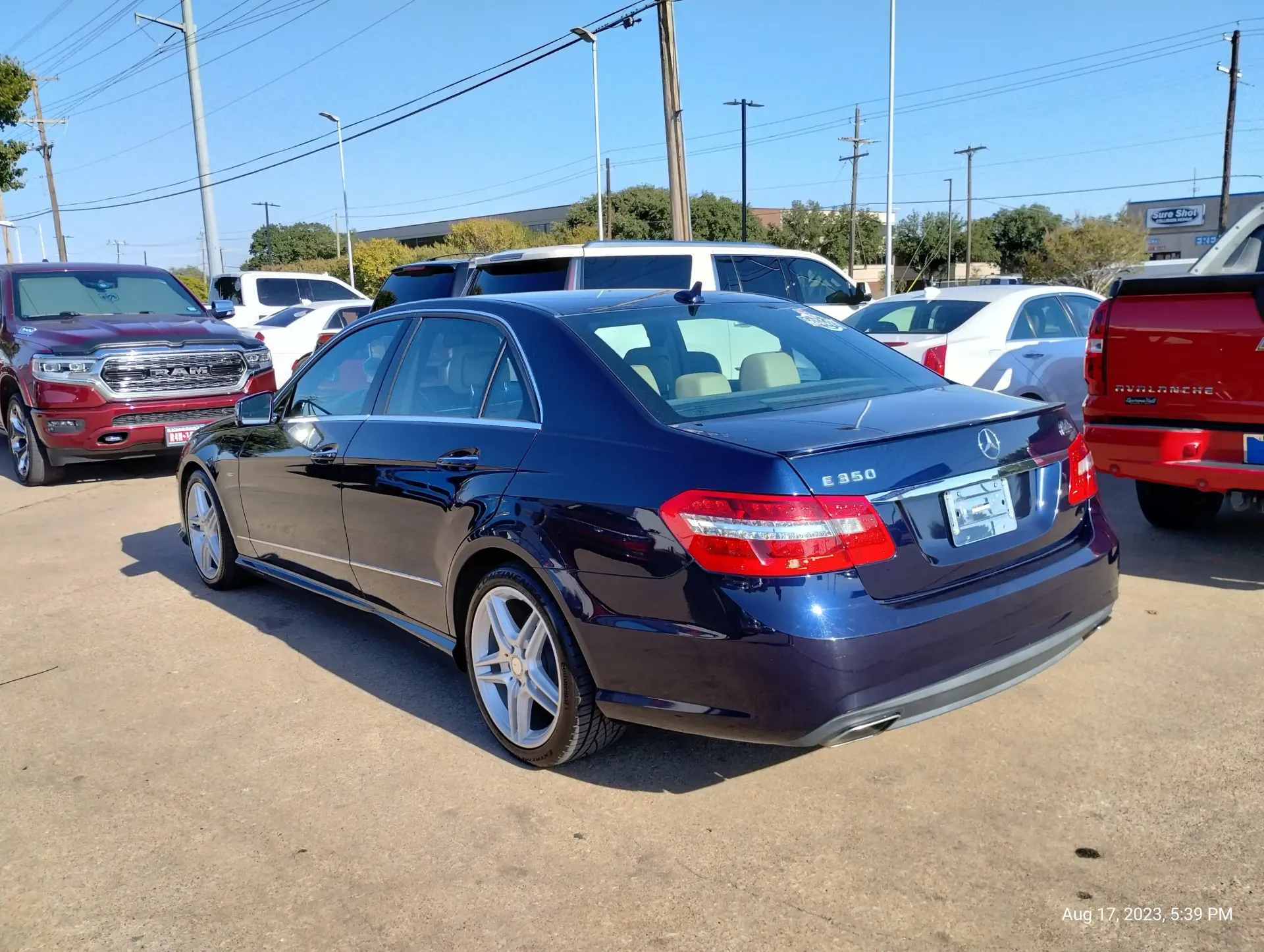 A dark blue Mercedes-Benz E-Class sedan parked on a gravel lot under a clear blue sky, surrounded by other vehicles.