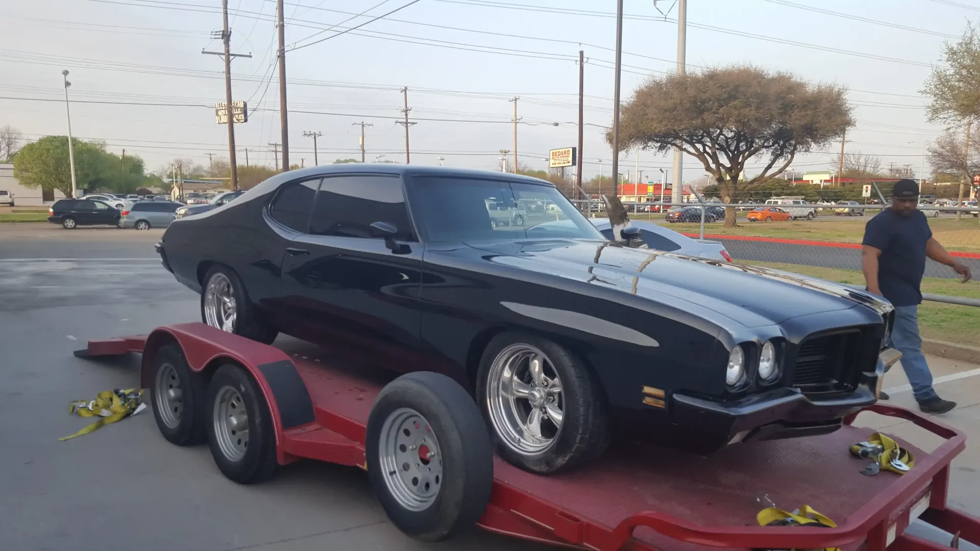 A black classic muscle car secured on a red flatbed trailer in a parking lot, with a person walking nearby.