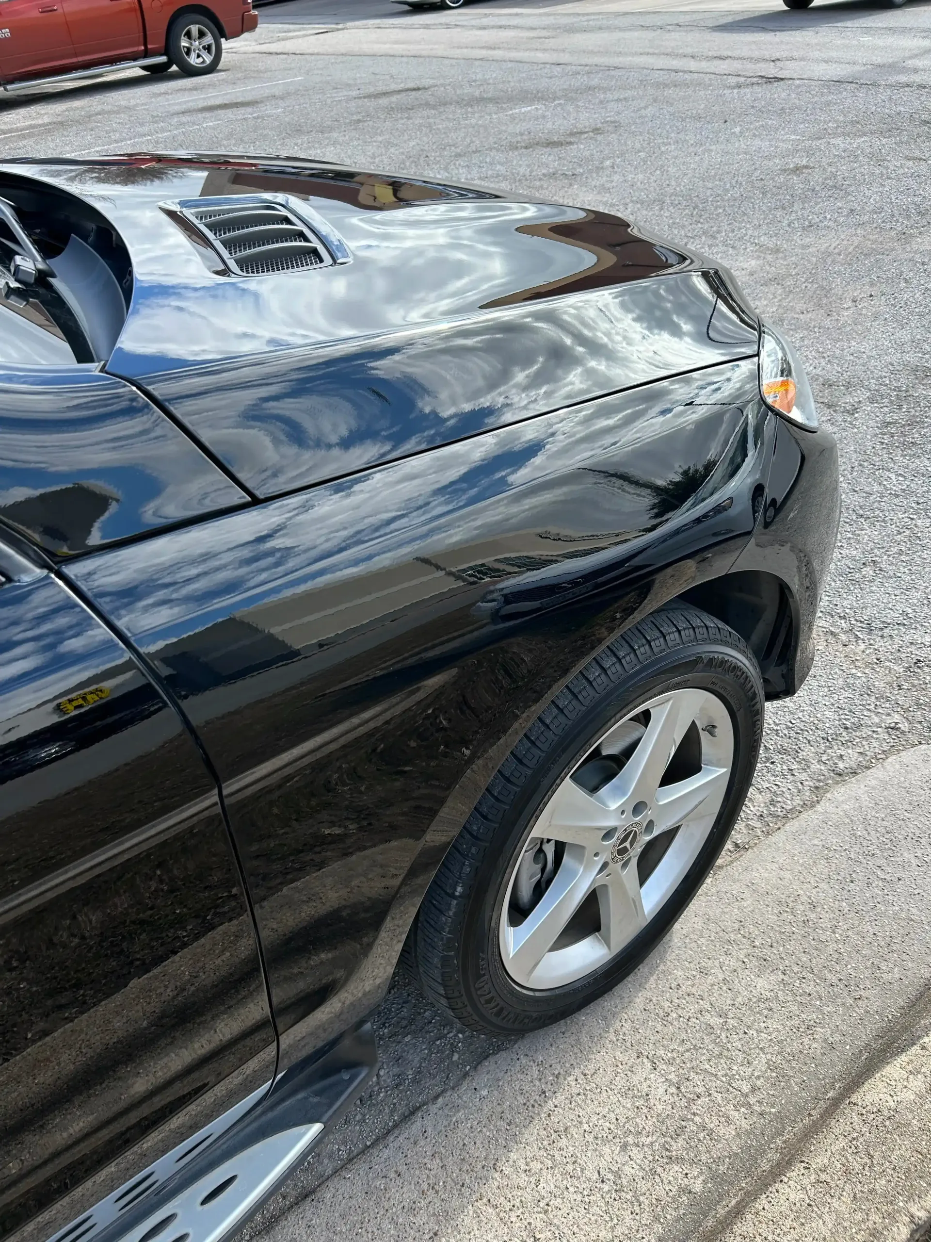 The front quarter view of a glossy black SUV parked on gravel, showing its hood, fender, and silver wheel.