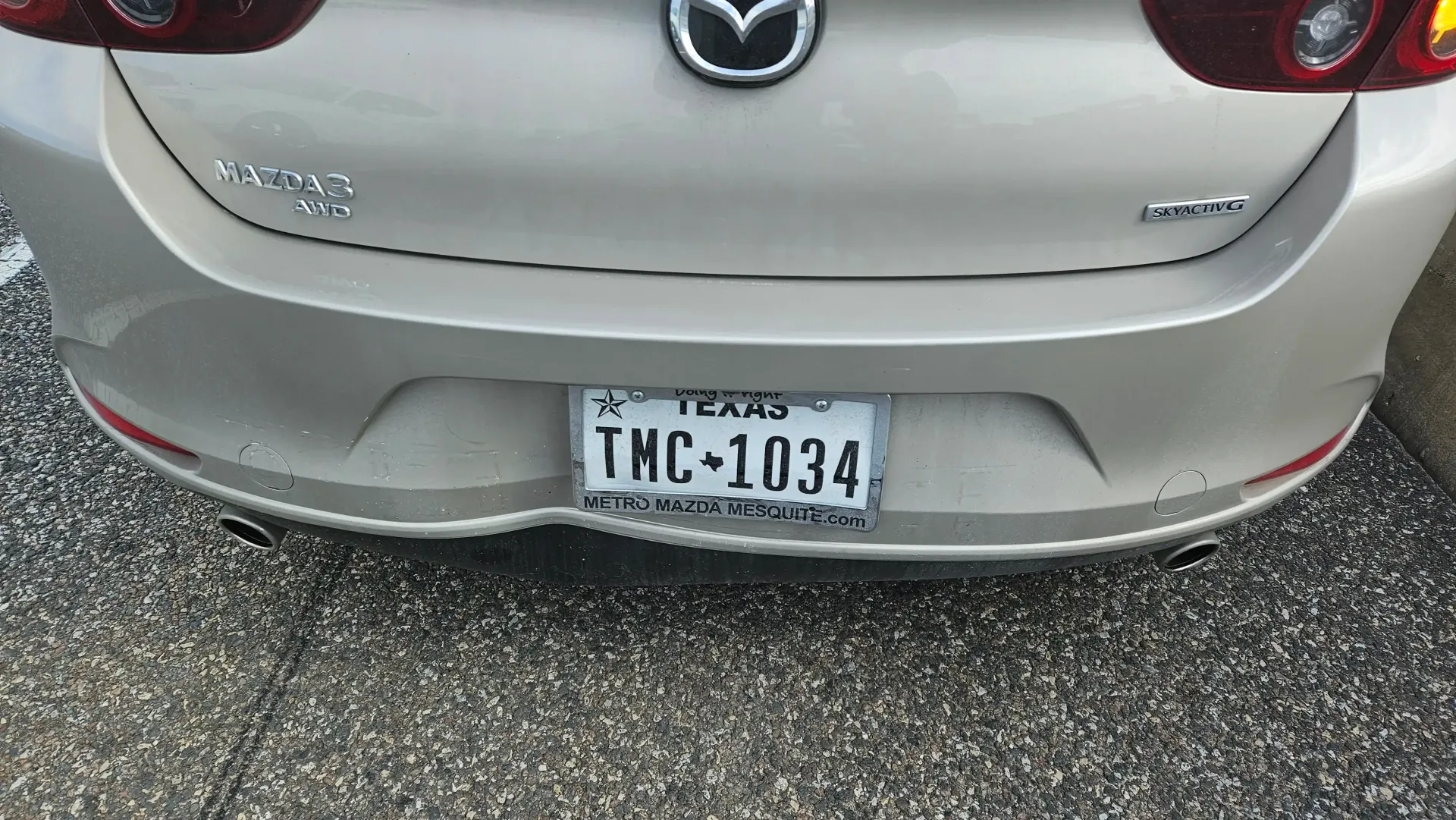 Rear view of a light-colored Mazda car with a Texas license plate reading TMC-1034 parked on asphalt.