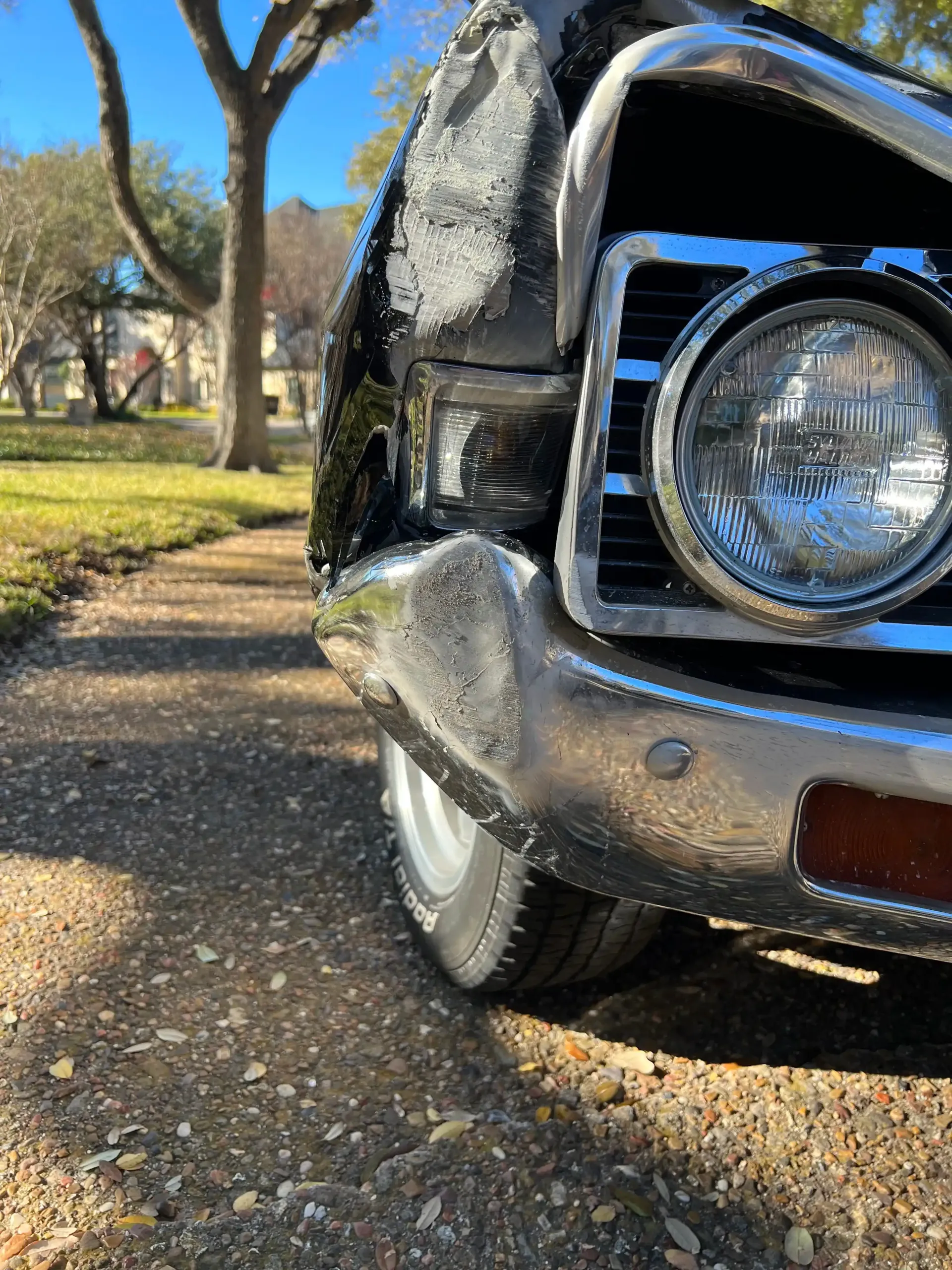 Close-up of a damaged front bumper and headlight of a classic black car parked on a gravel path near grass and trees.