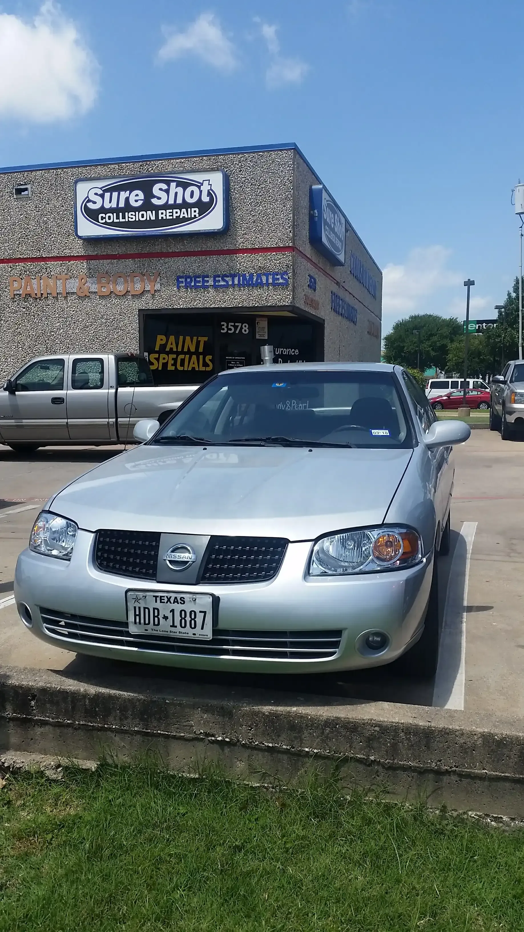 A silver Nissan sedan parked in front of a Sure Shot business building under a blue, cloudy sky.