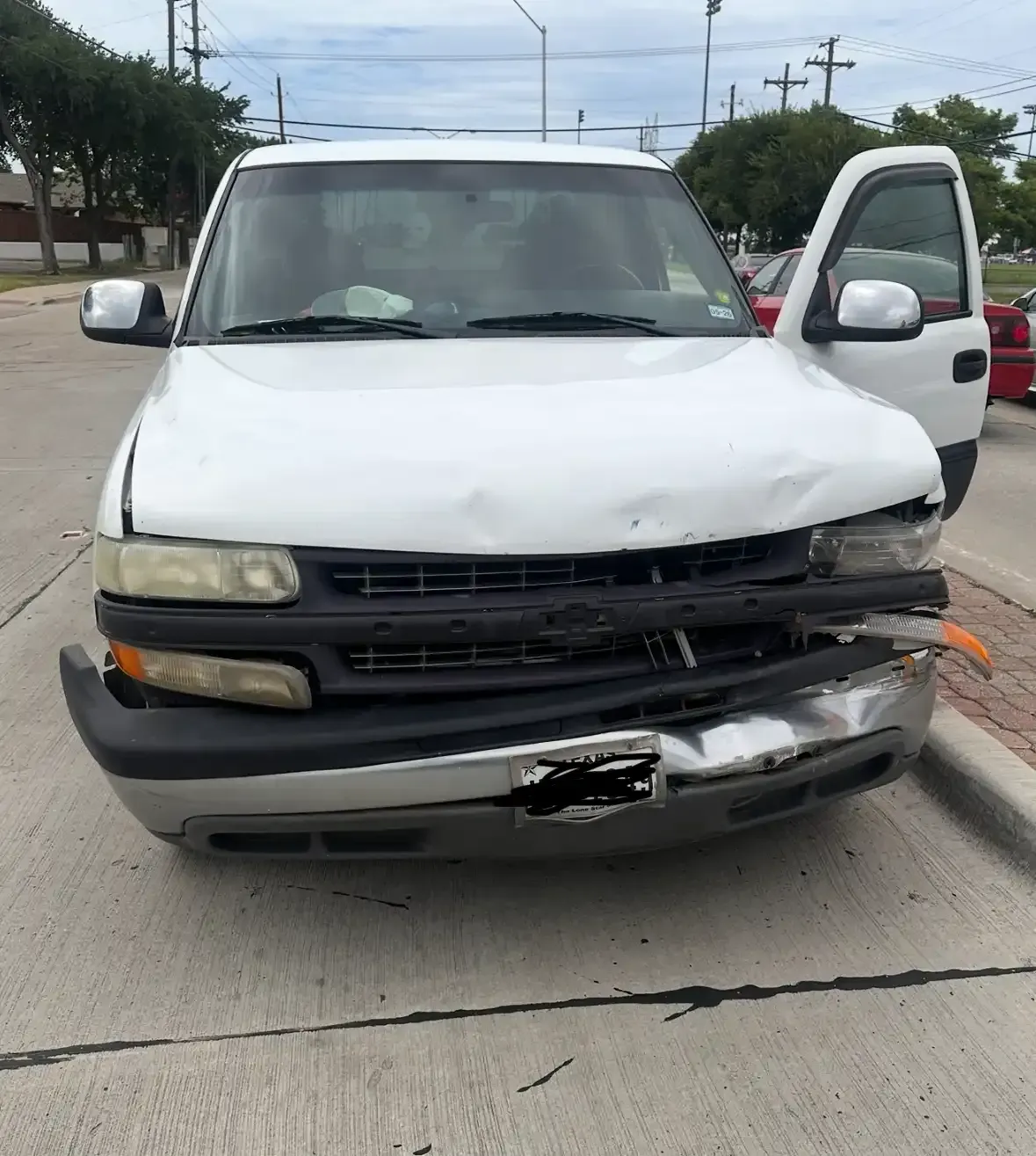 A white pickup truck parked on a street with significant front-end collision damage to the bumper, grille, and hood.