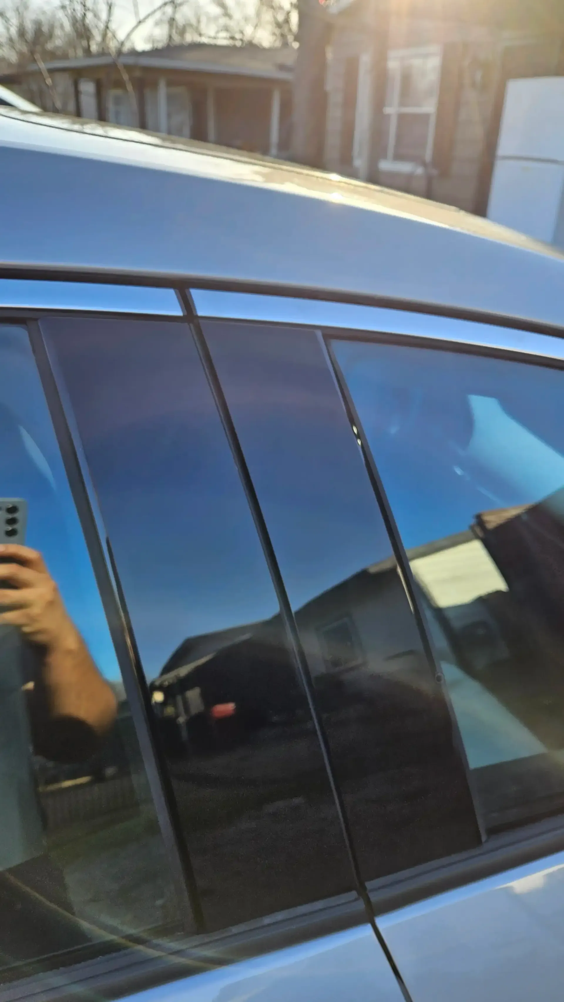 Close-up of a car's black glossy pillar trim between the front and rear windows, reflecting the sky and a house.