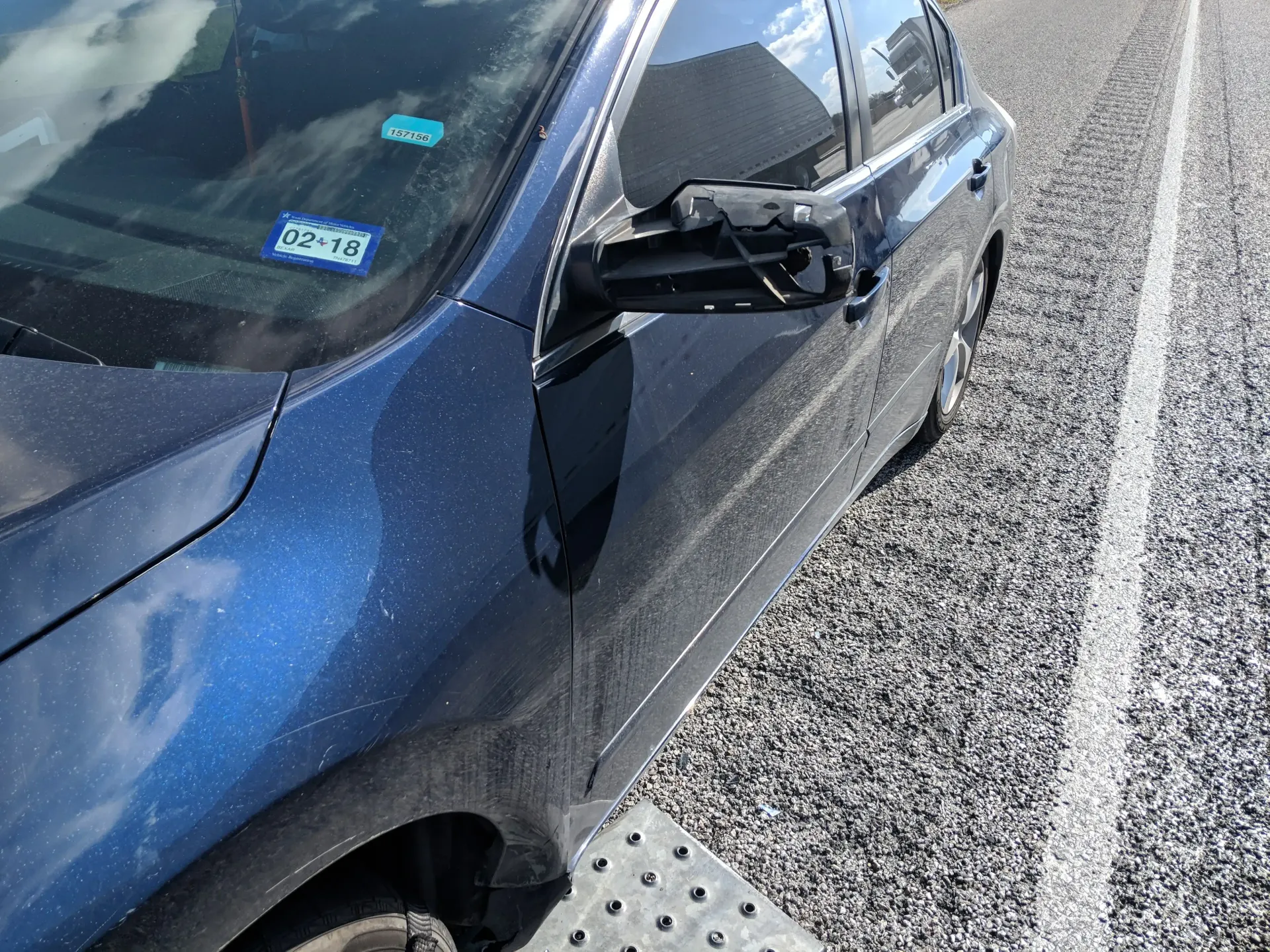 A side-view of a blue car parked on gravel, showing a damaged side mirror with the housing missing.