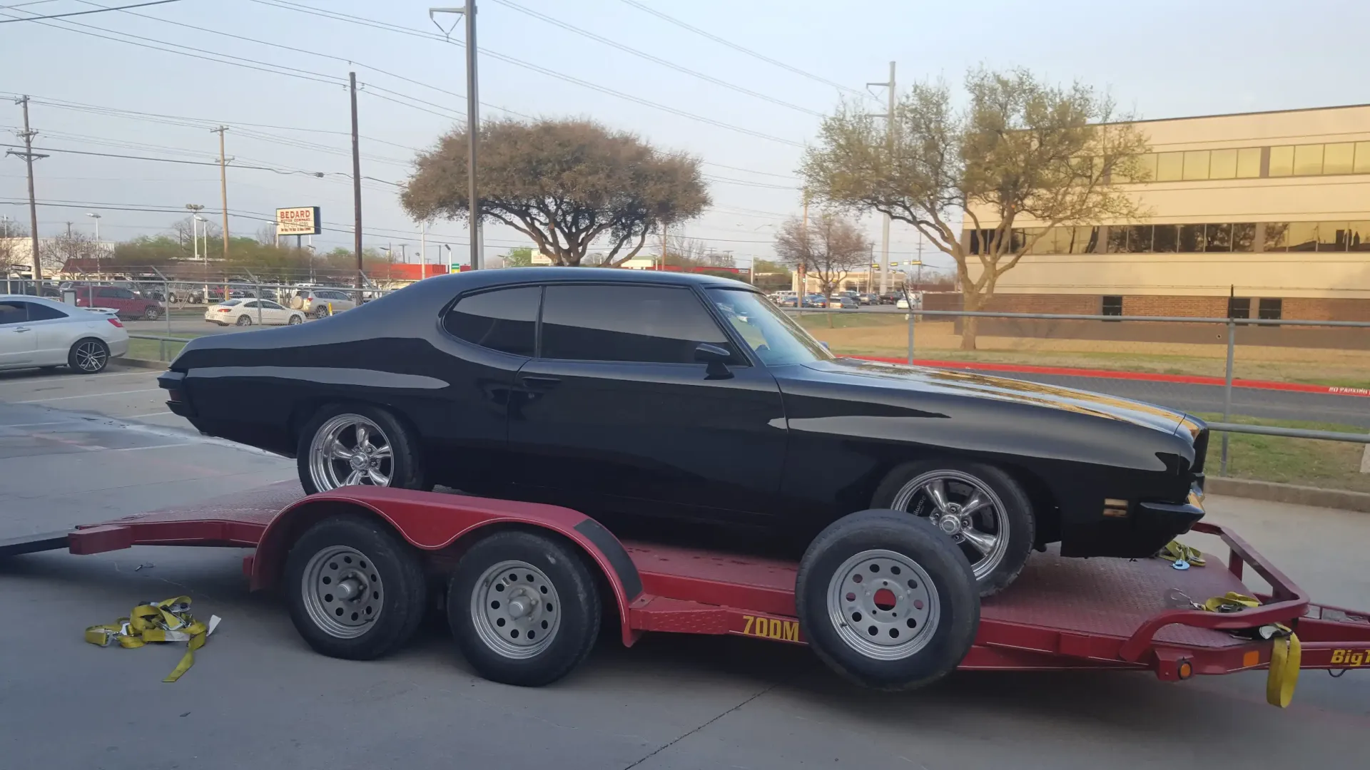 A glossy black vintage muscle car secured on a red flatbed car hauler in a paved parking lot.