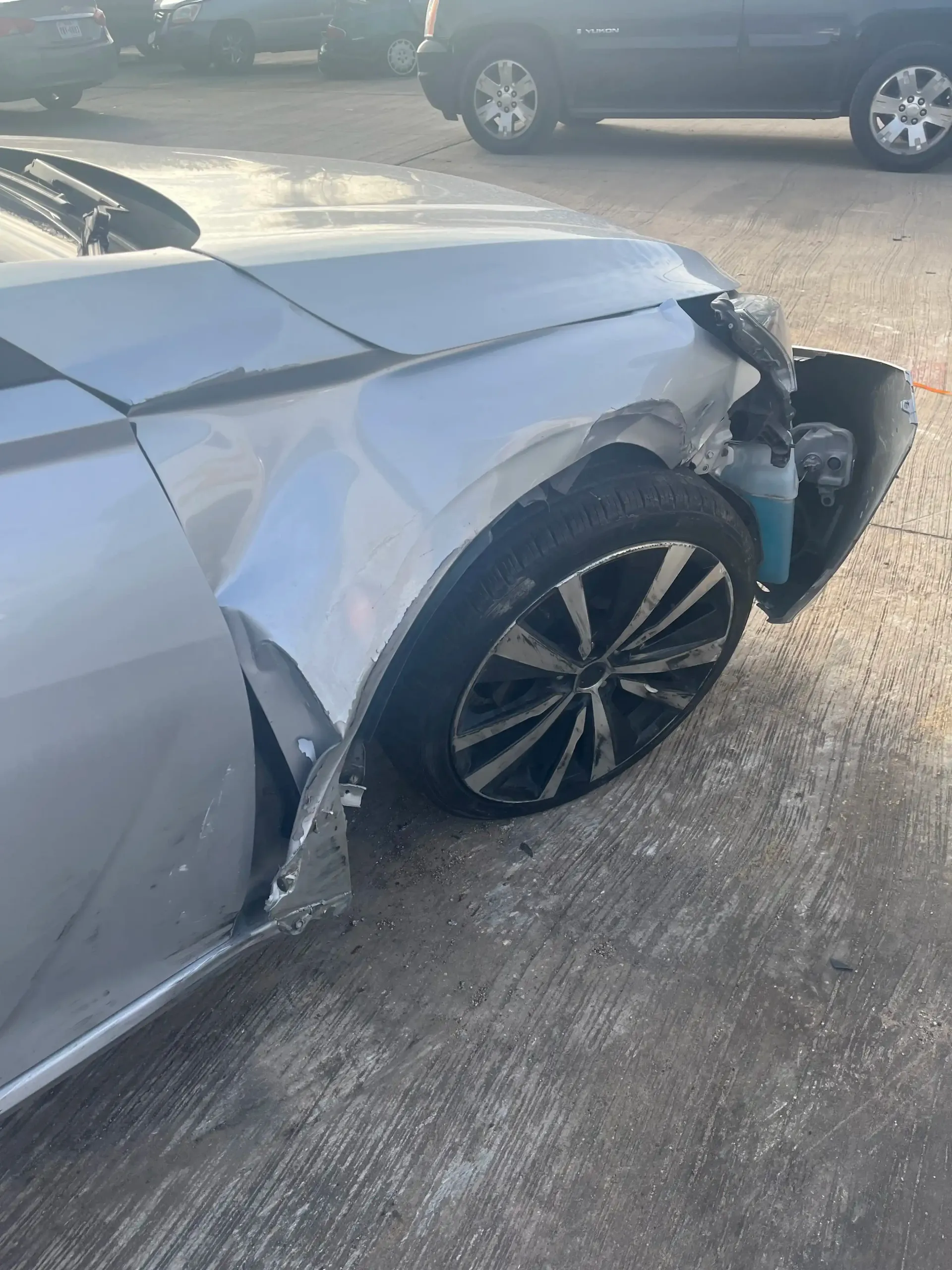 Silver car with significant front-end damage, including a crumpled fender, missing bumper, and damaged wheel well.