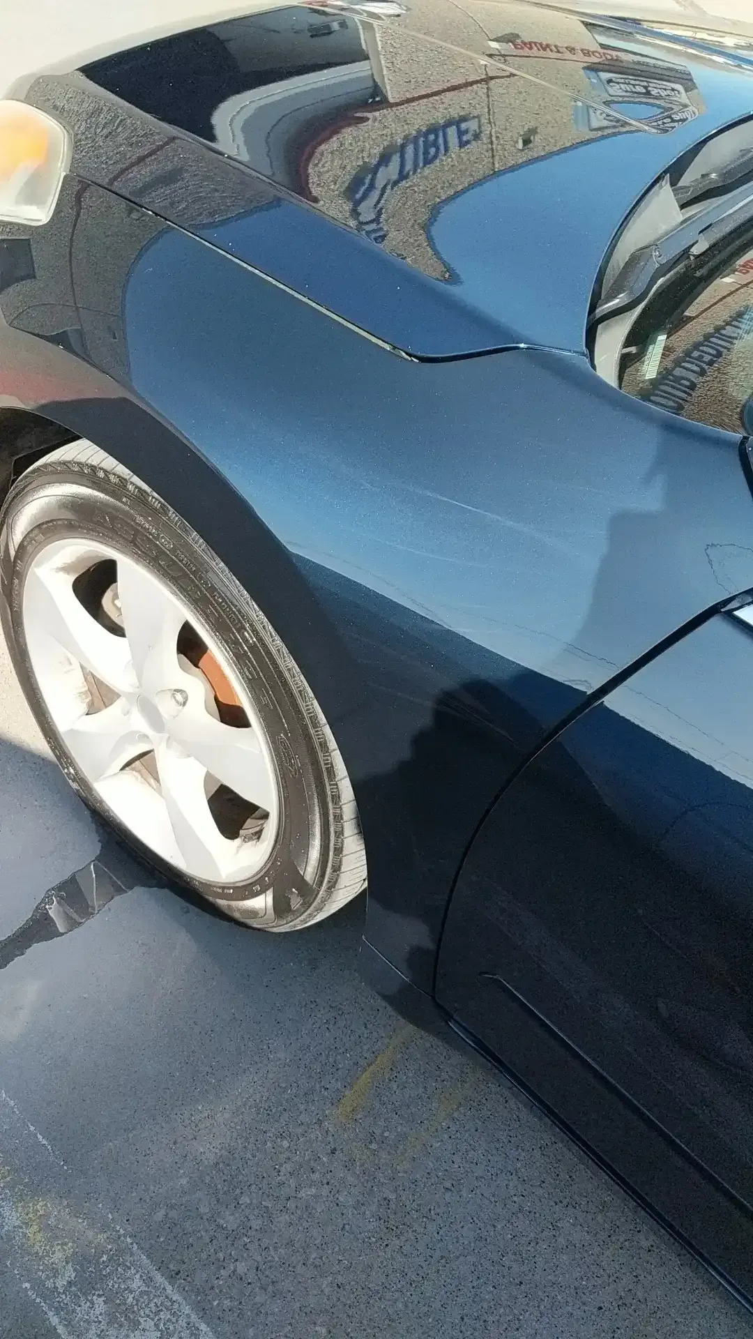 A close-up of a dark blue car's front fender and wheel, parked on gray pavement.