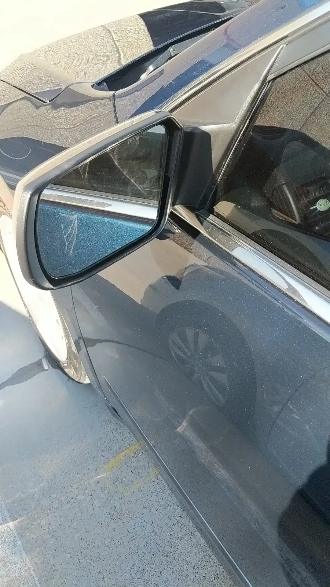 A close-up of a side mirror on a dark grey car, showing its reflection in bright outdoor lighting.
