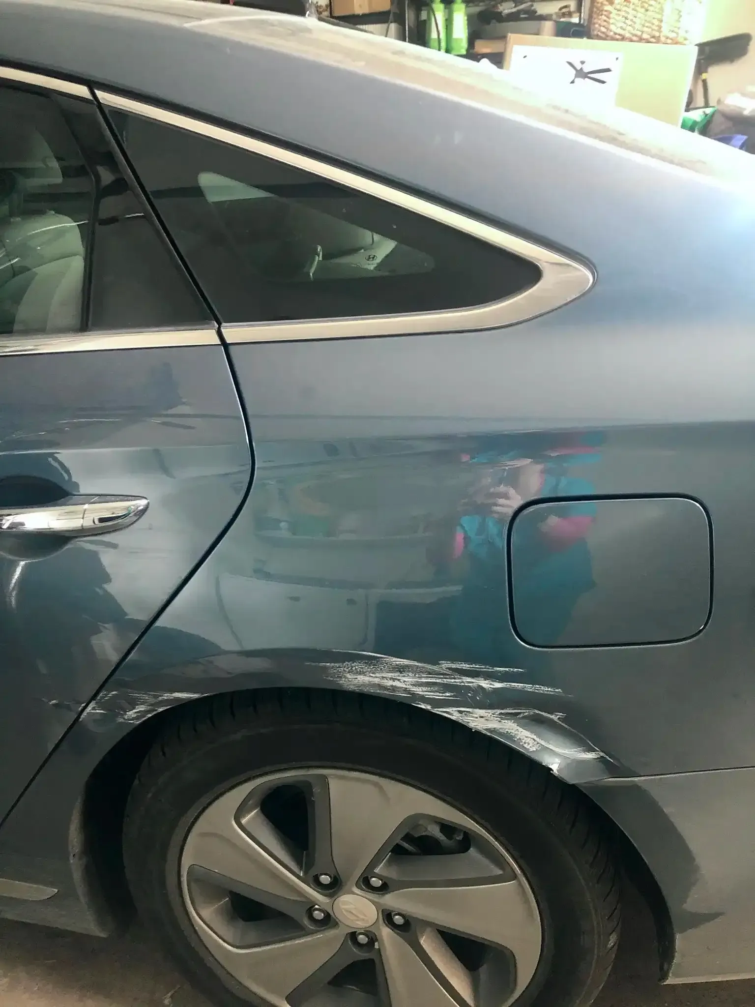 A close-up view of the rear side panel of a blue car showing significant paint damage and scraping above the rear wheel.
