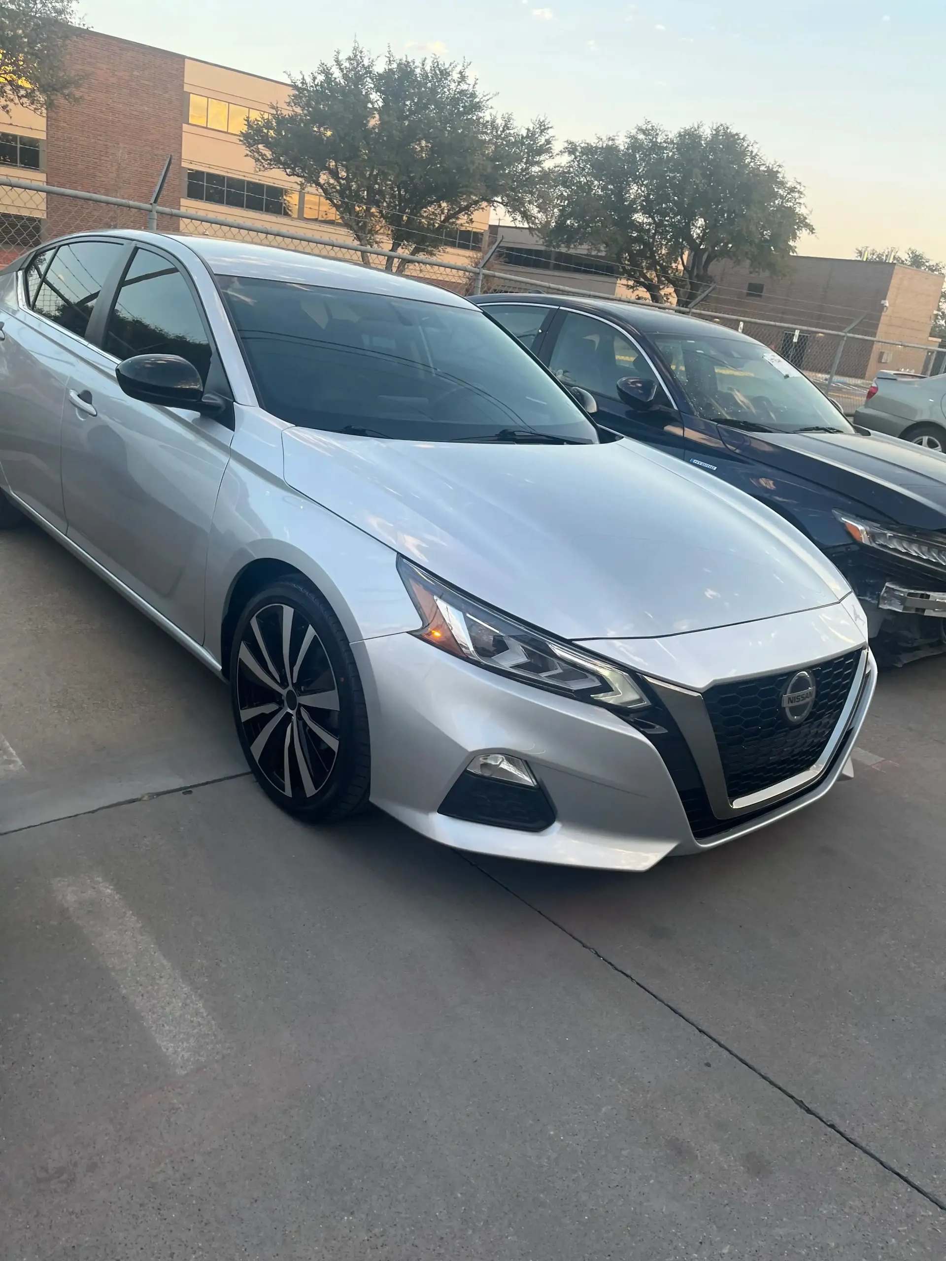 A silver Nissan Altima parked in a paved lot during the day, with another vehicle parked beside it.