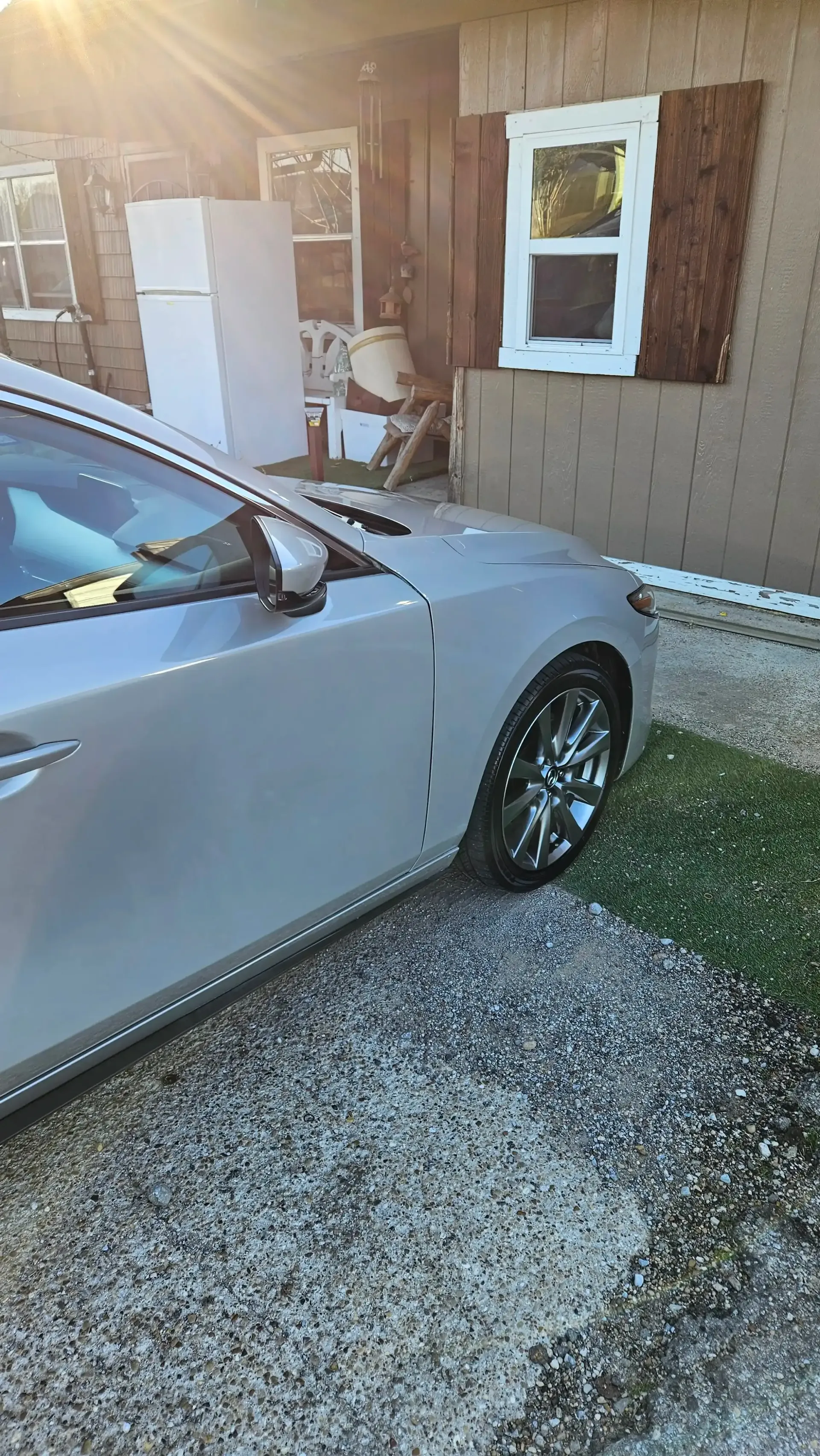 A silver car parked on a gravel driveway in front of a house with a white refrigerator visible on a porch.