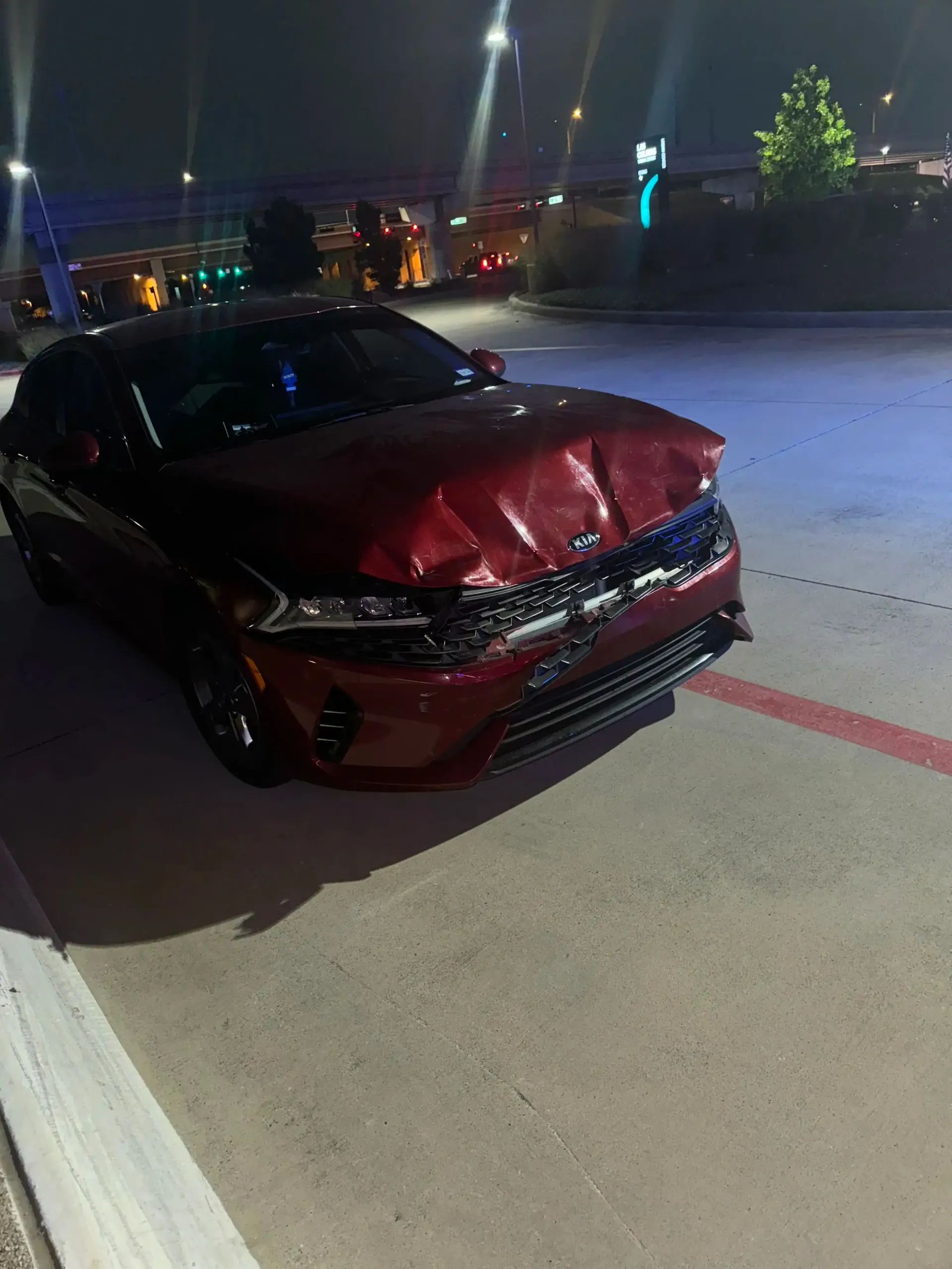 A dark red sedan parked at night with severe front-end damage, including a crumpled hood and damaged front grille.