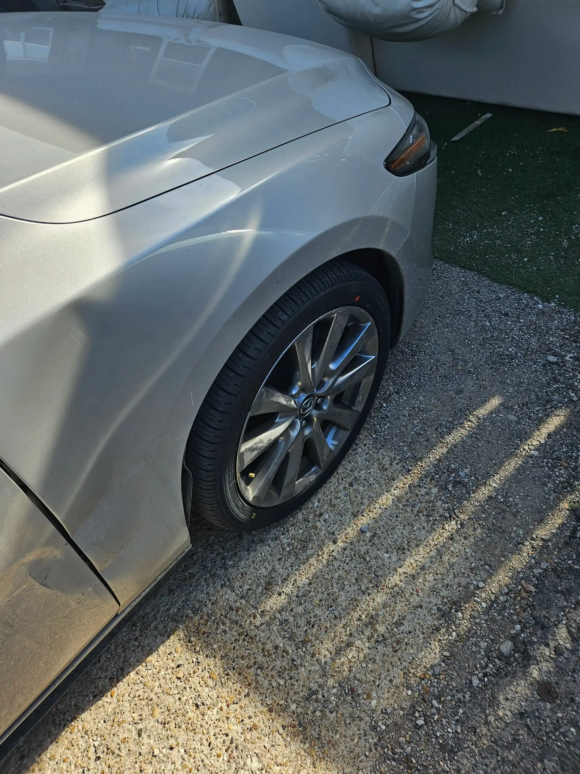 A silver car parked on a gravel driveway, showing the front passenger-side wheel and headlight.