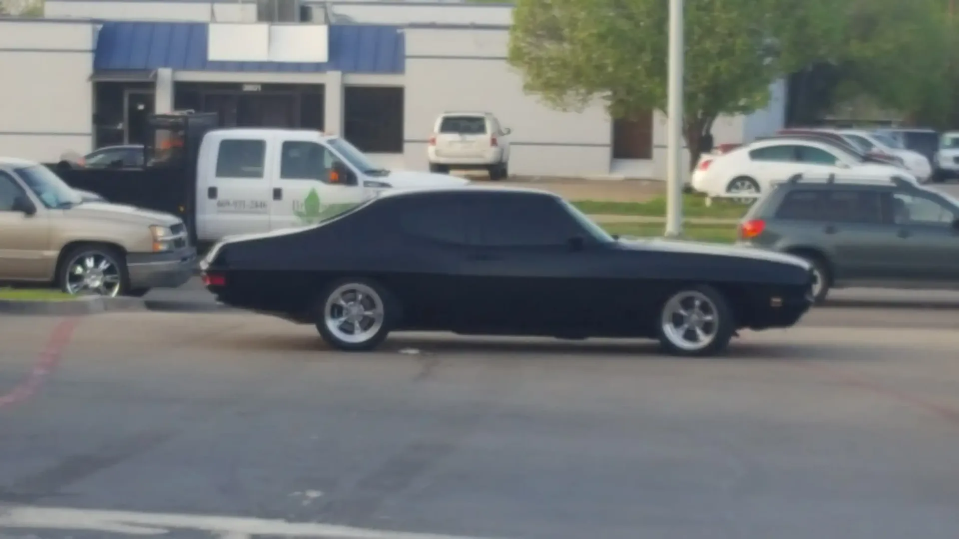 A black vintage muscle car driving across an asphalt parking lot in front of commercial buildings and parked vehicles.