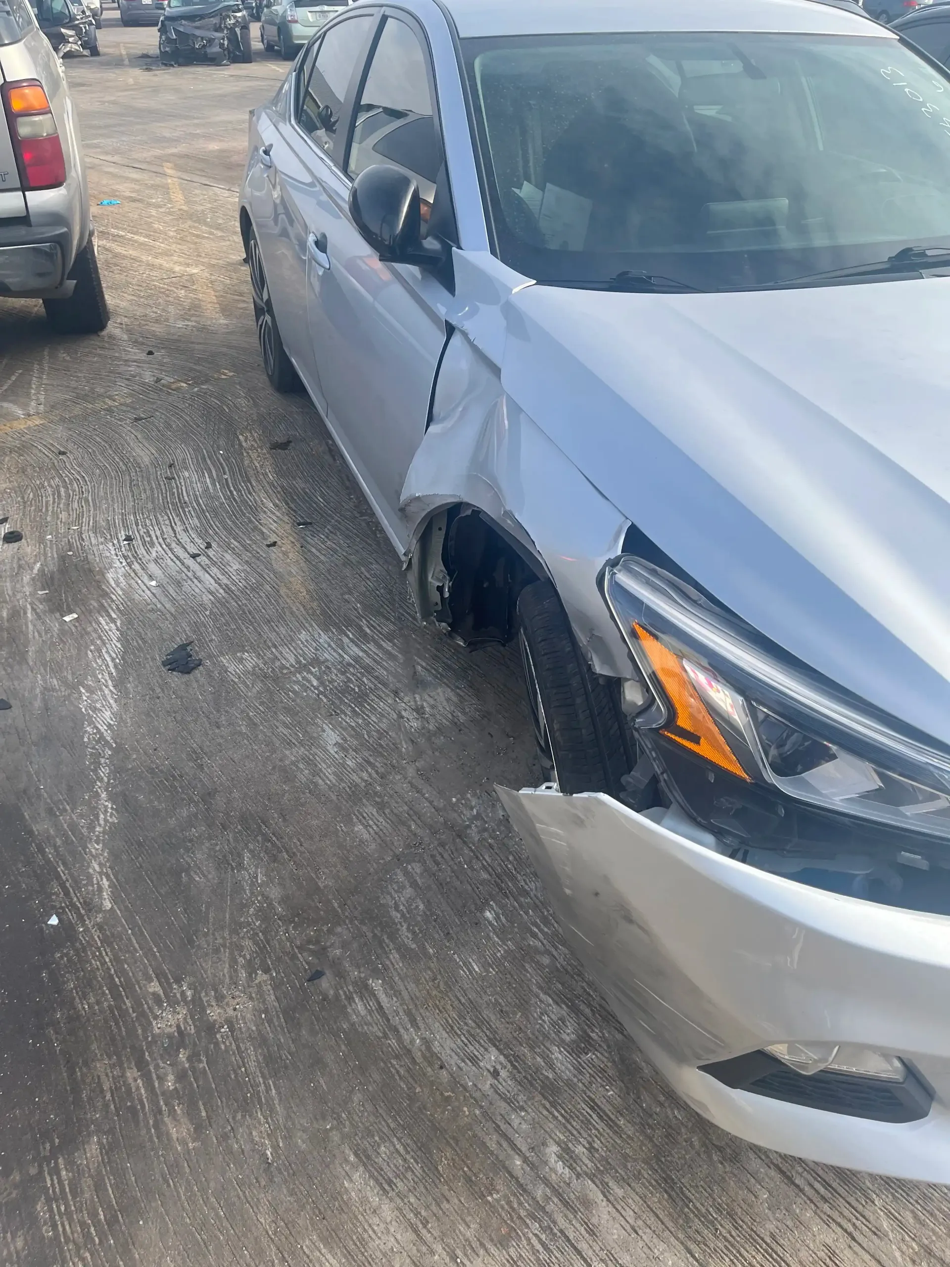 A silver car parked on asphalt with significant damage to the front bumper and front passenger-side fender.