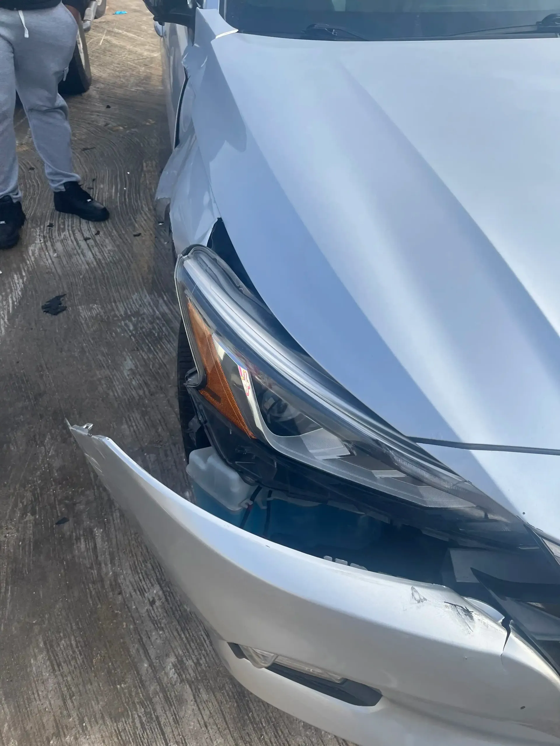 A silver car with damage to its front bumper and headlight, detached from the vehicle's side, on a paved surface.