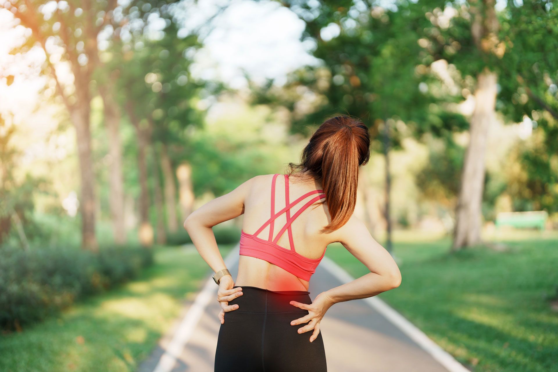 Woman in activewear clutches back, standing on path in park, likely experiencing back pain.
