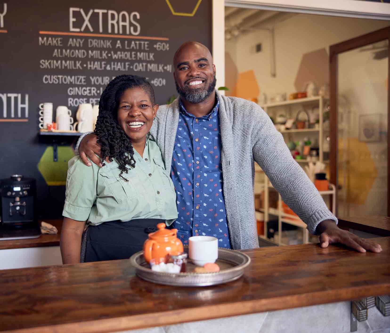 Two people standing behind a cafe counter, smiling. Coffee and tea items are on a tray.