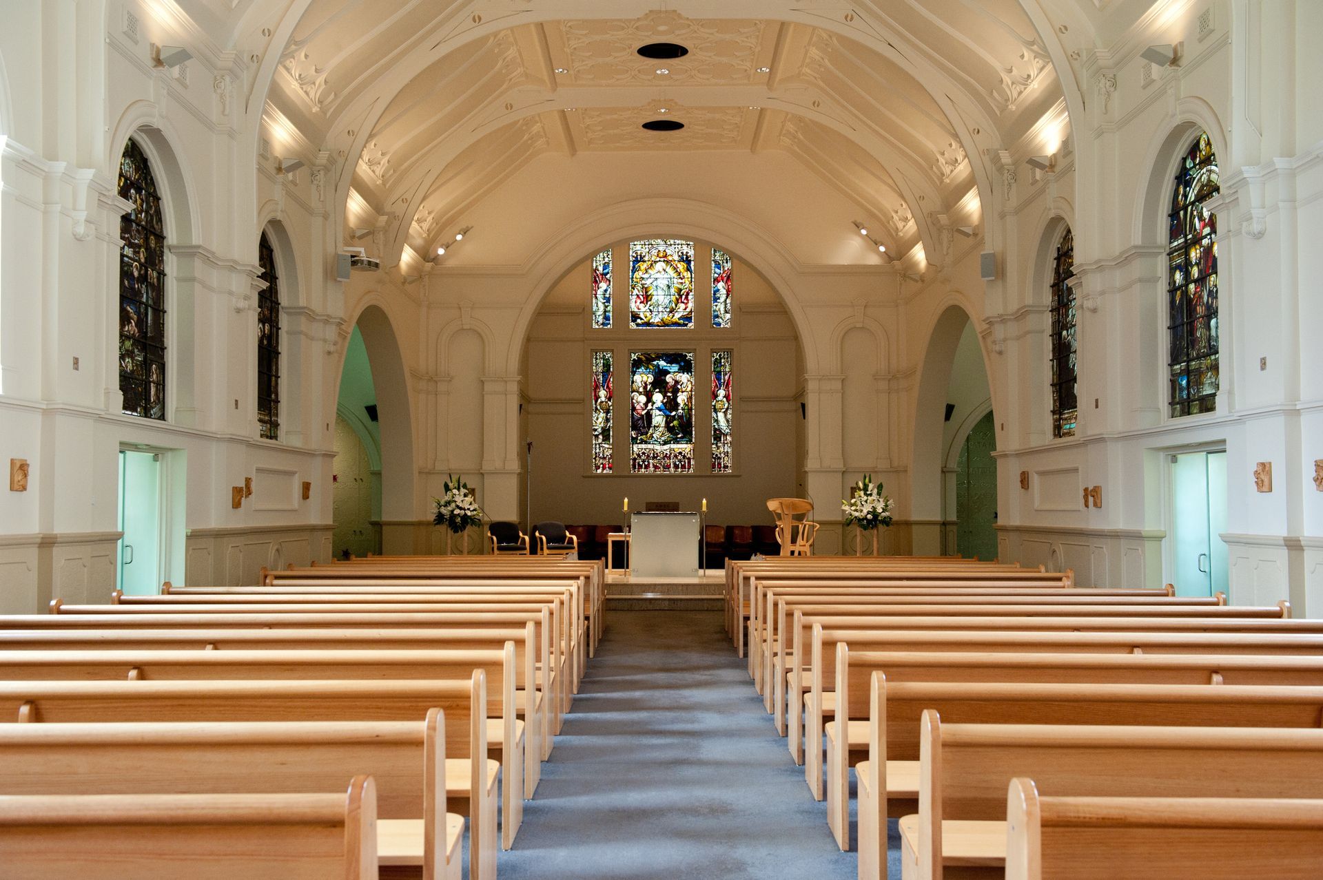 Interior of a church or chapel with rows of wooden pews leading to a central altar and stained-glass windows.