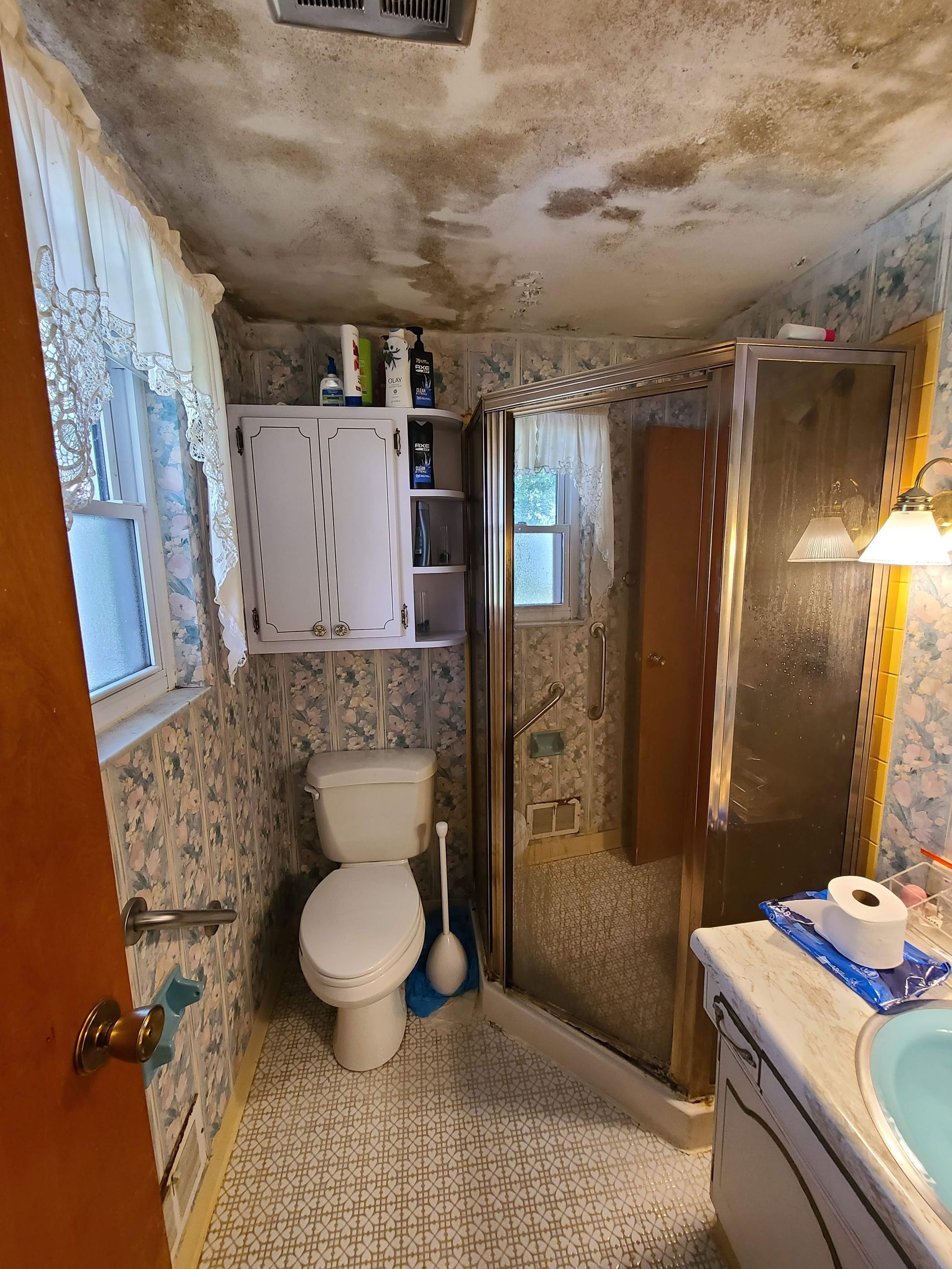 Small bathroom with mold on the ceiling, toilet, shower, vanity, and patterned wallpaper.