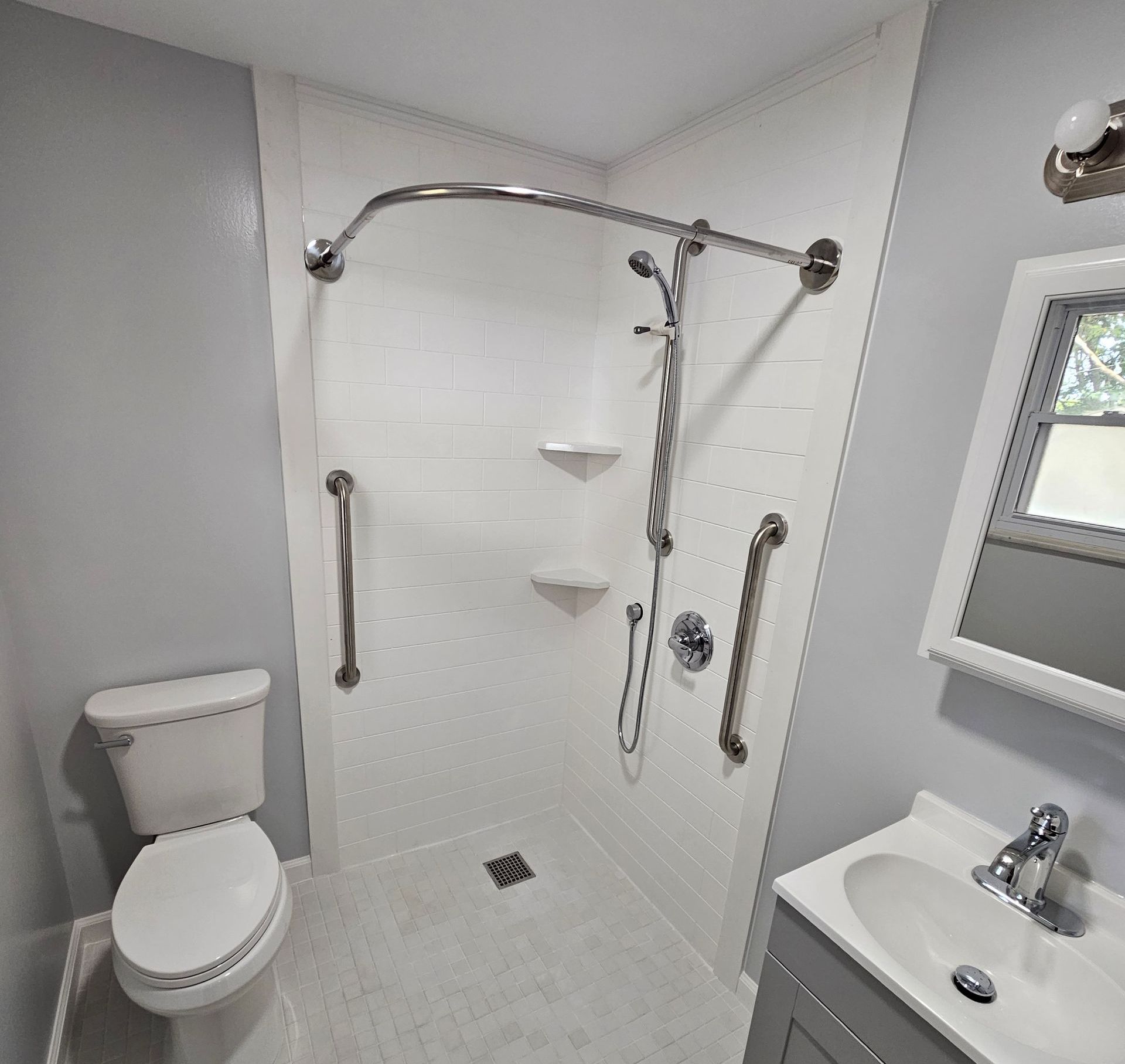 Bathroom with walk-in shower, toilet, and vanity. White shower walls, silver grab bars, gray walls, and fixtures.