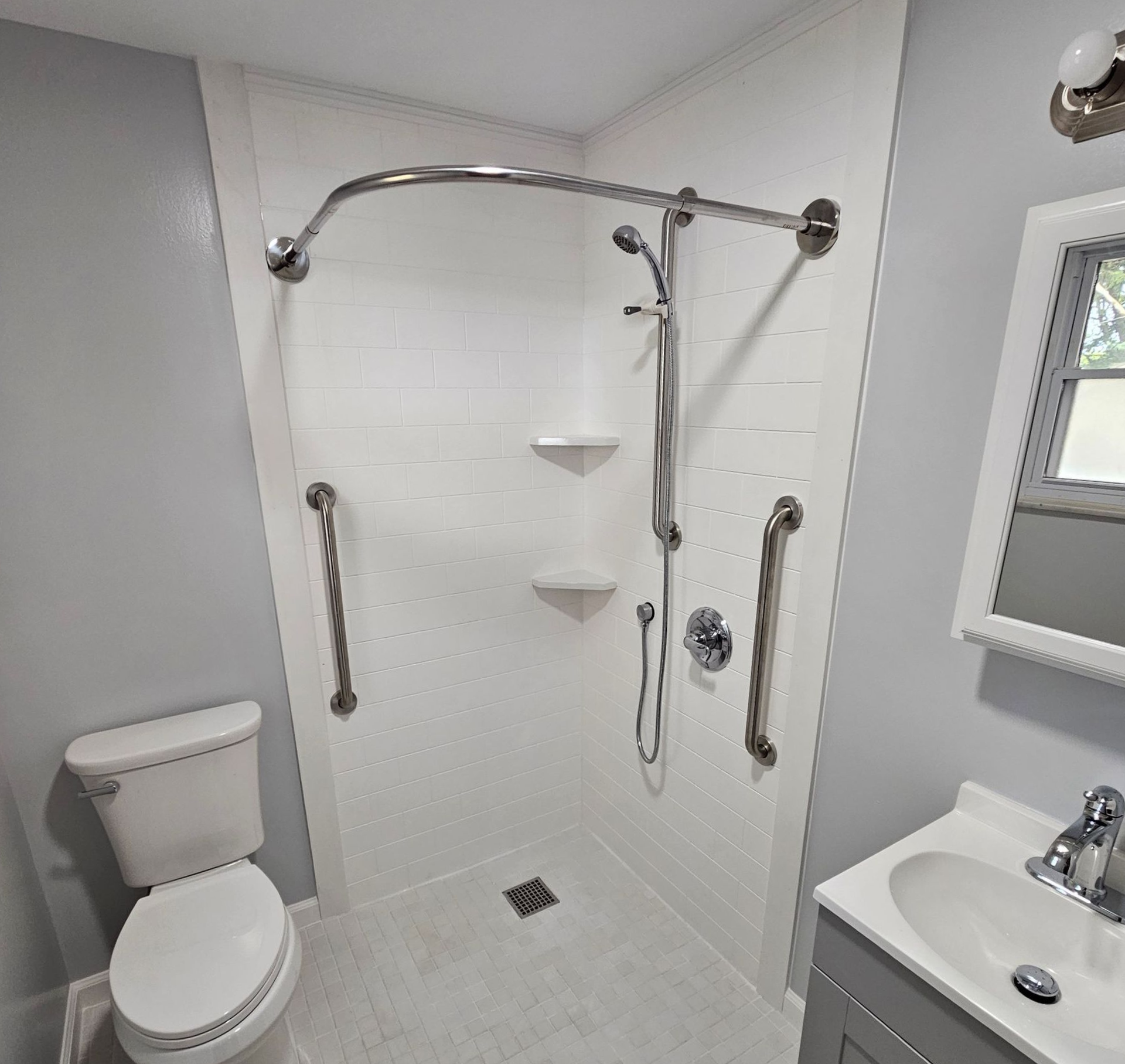 Accessible bathroom with white shower, grab bars, toilet, sink, and pale gray walls.