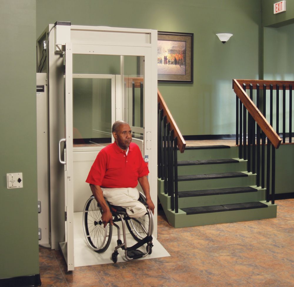 Man in wheelchair exits residential lift next to stairs; green walls, painting, and dark brown railing.