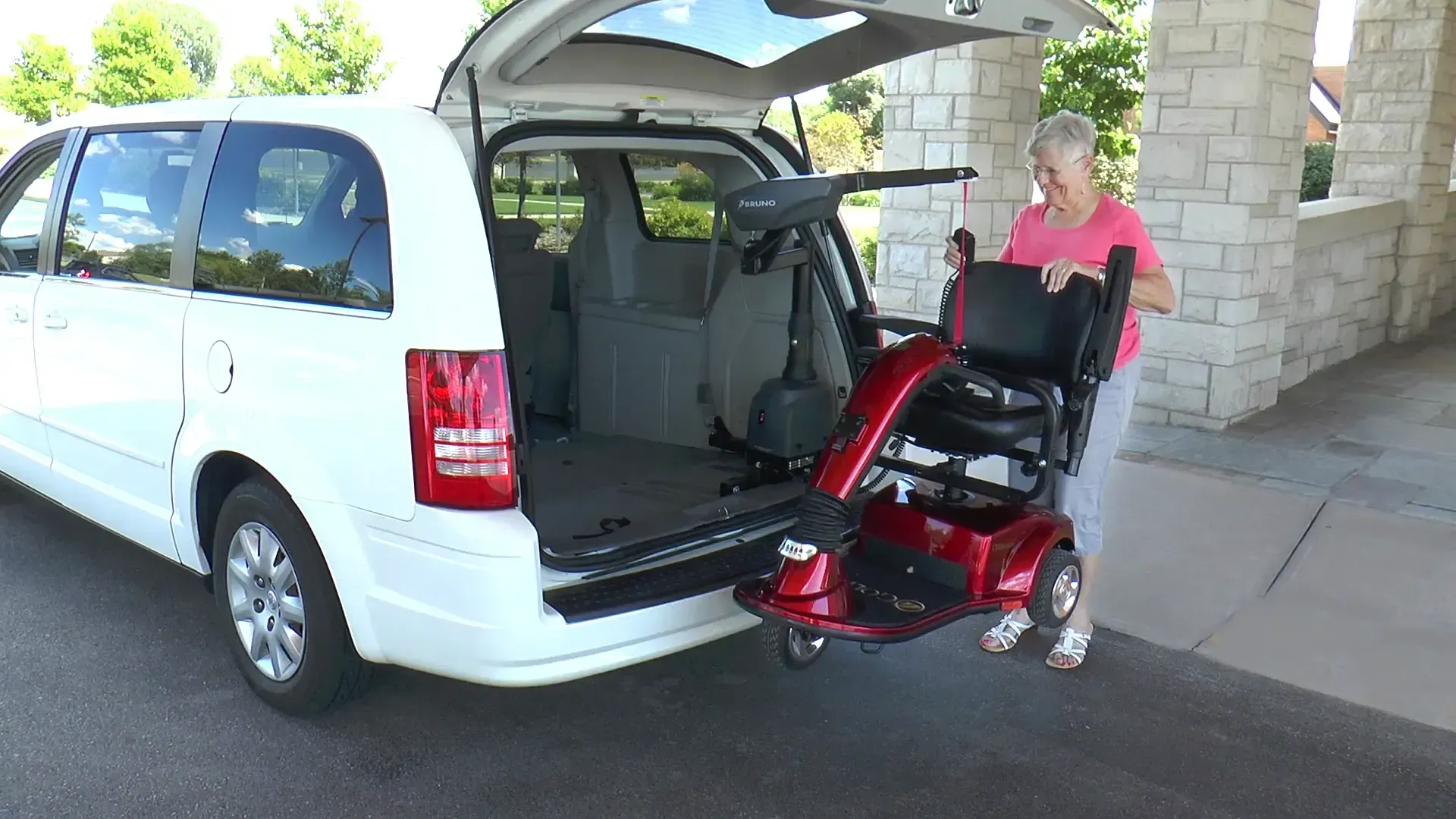Person loading a red mobility scooter into the back of a white minivan in a driveway