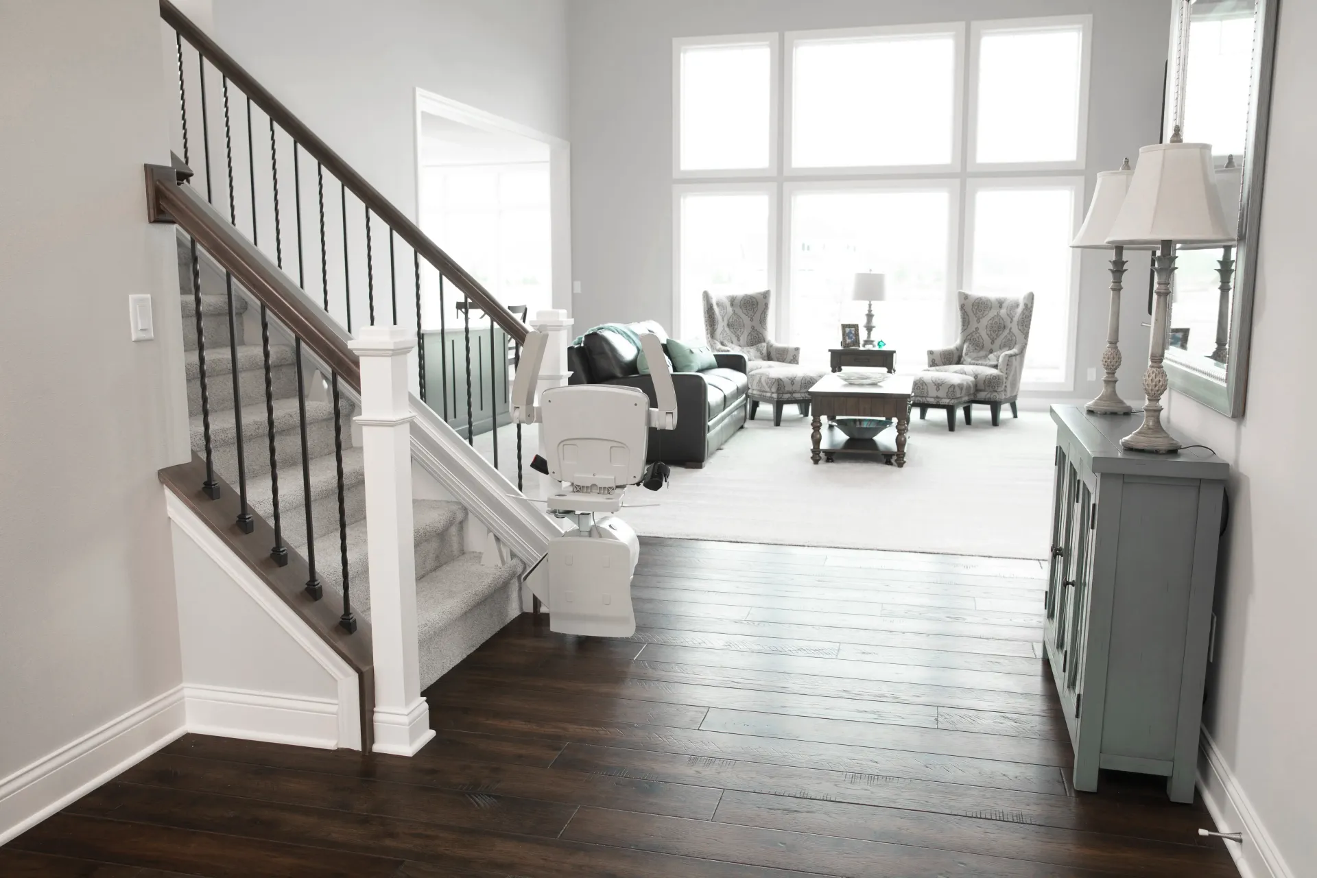 Bright, airy living room with dark wood floors, white stairs, large windows, and teal furniture.