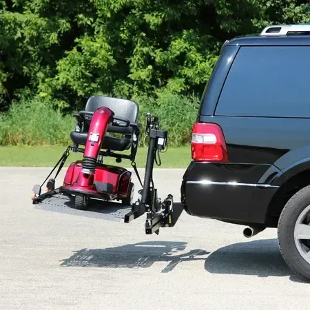 A red mobility scooter on a lift attached to the back of a black SUV. Outdoors, green trees.