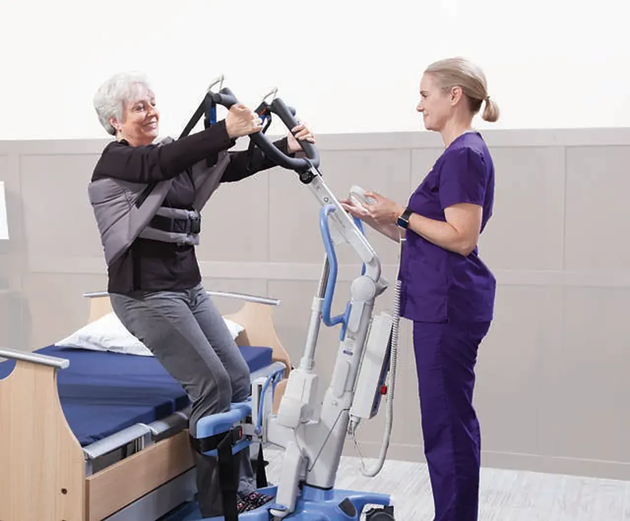Caregiver using a Hoyer Elevate sit-to-stand lift to raise a woman from the edge of a bed