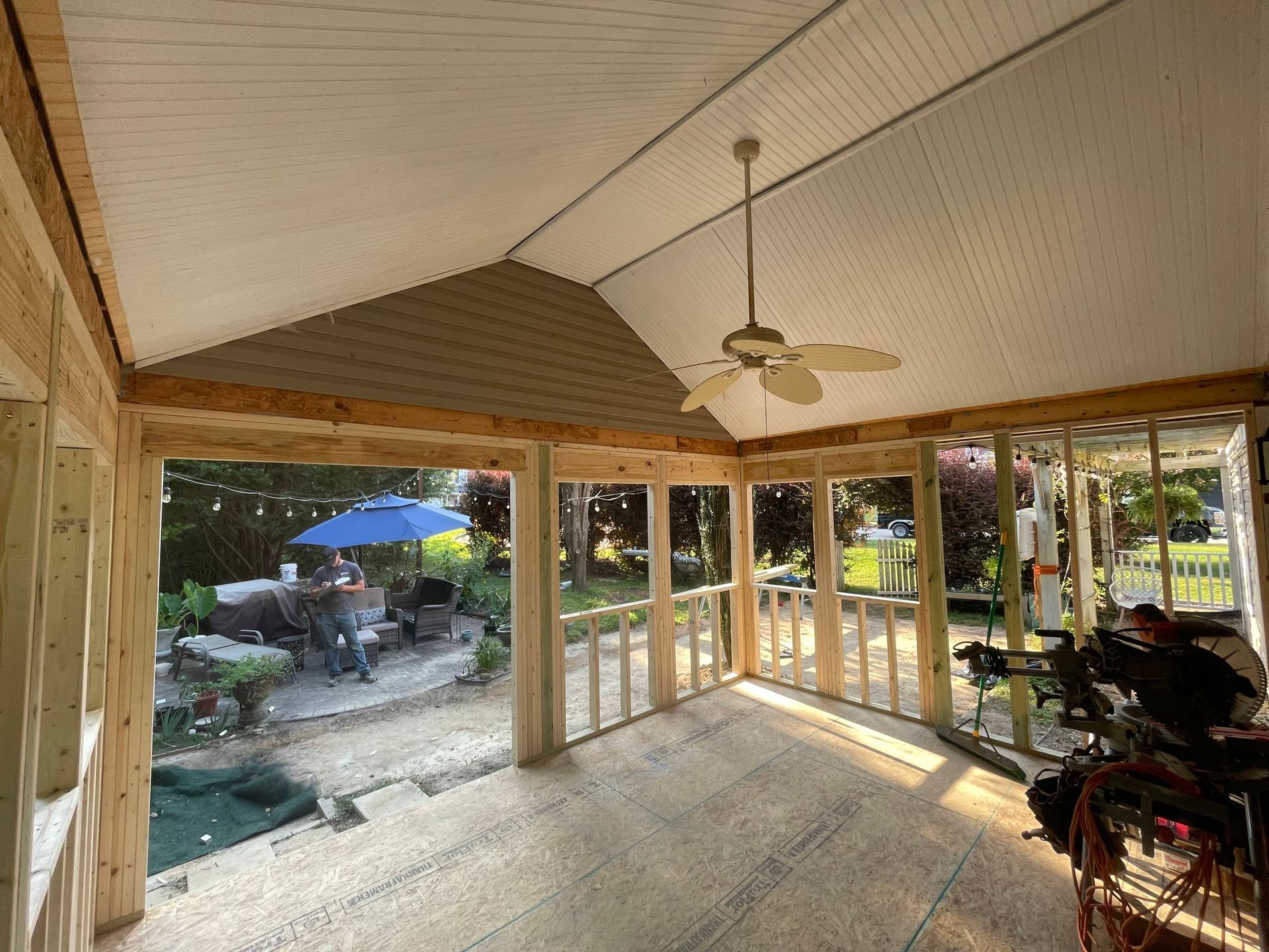 Interior view of a sunroom under construction, with exposed wood framing and a view of a backyard with patio furniture.