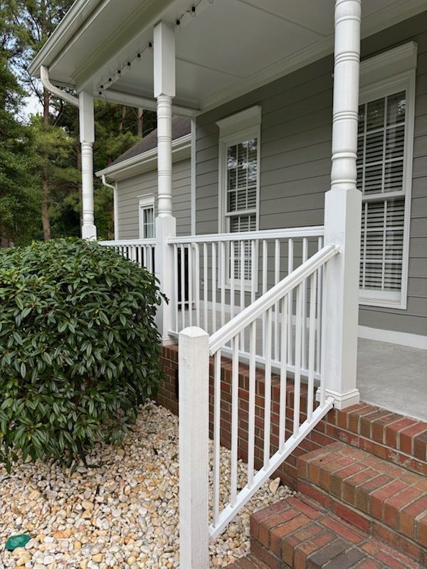 Front porch with white railing, brick steps, and gray house siding beside shrubs and gravel