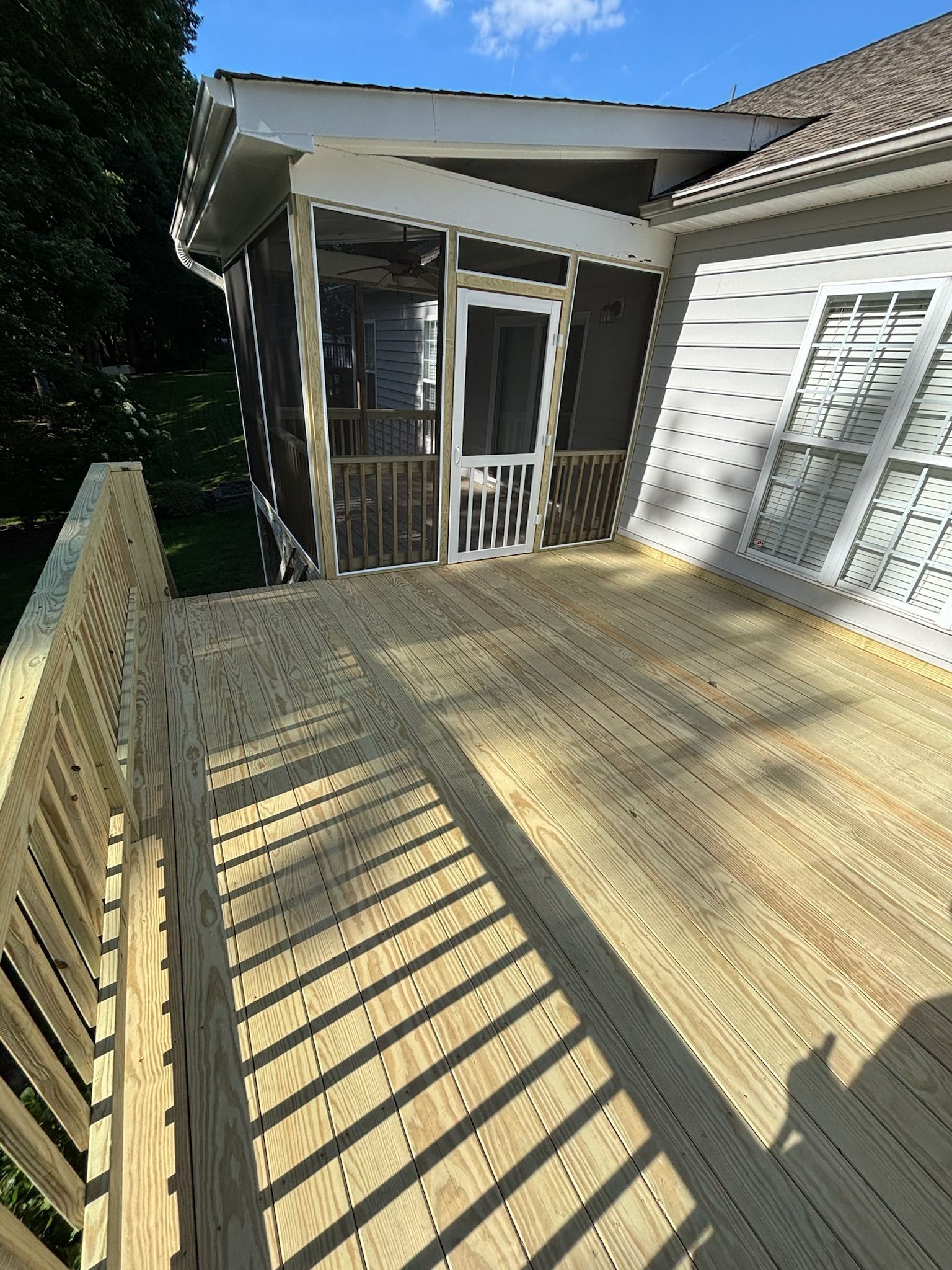 Sunlit wooden deck beside a white house, with railing shadows and screened porch entry