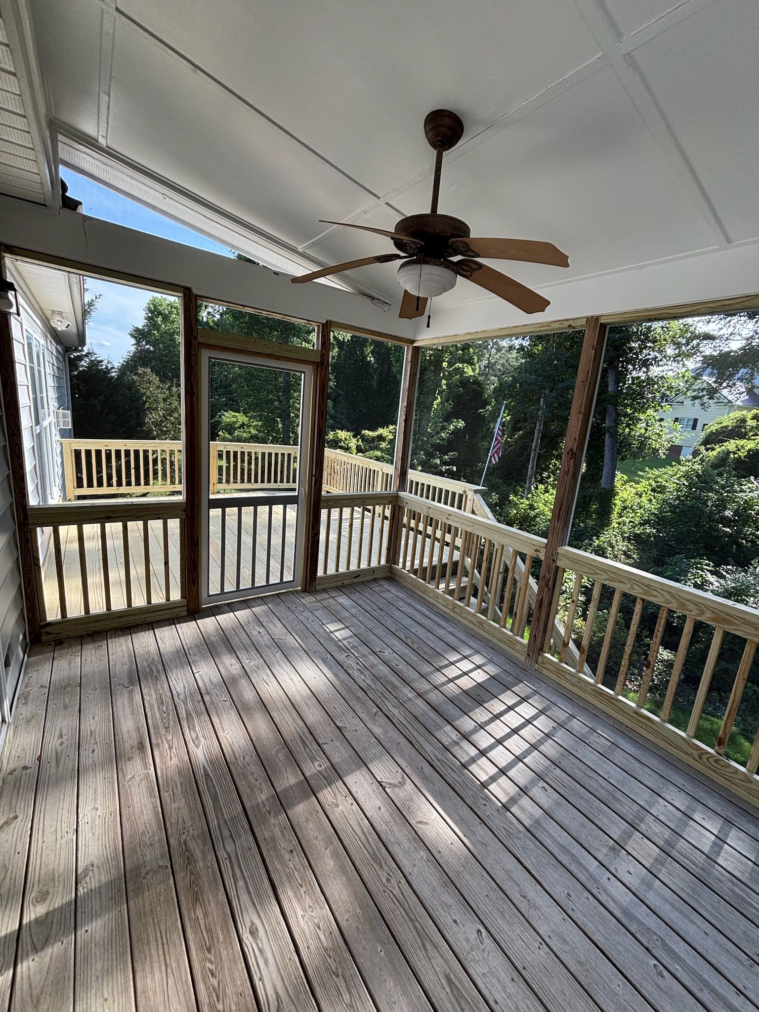 Covered wooden porch with railings, ceiling fan, and sunlight through trees.