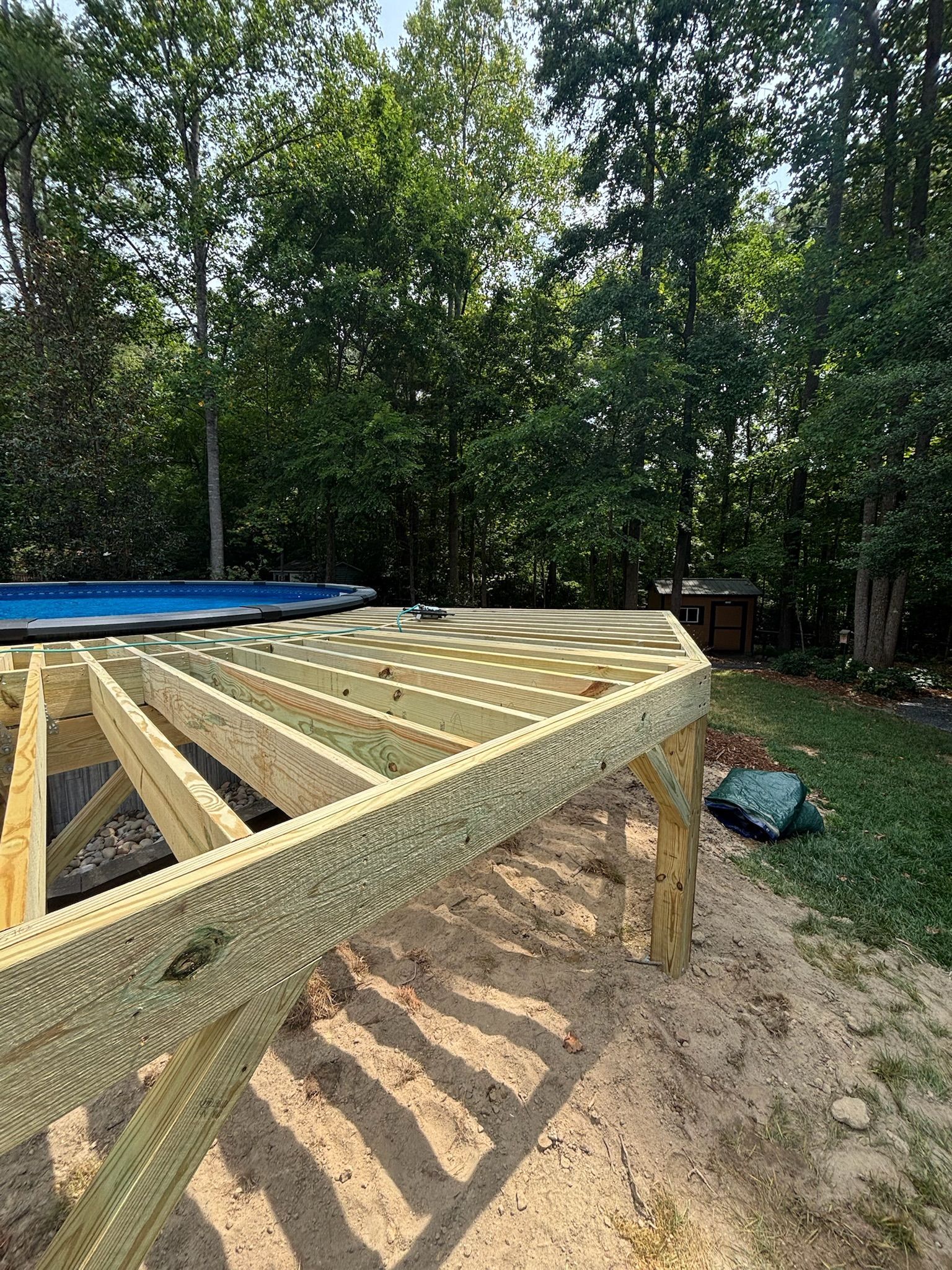 Wooden deck frame under construction beside a blue above-ground pool in a wooded backyard