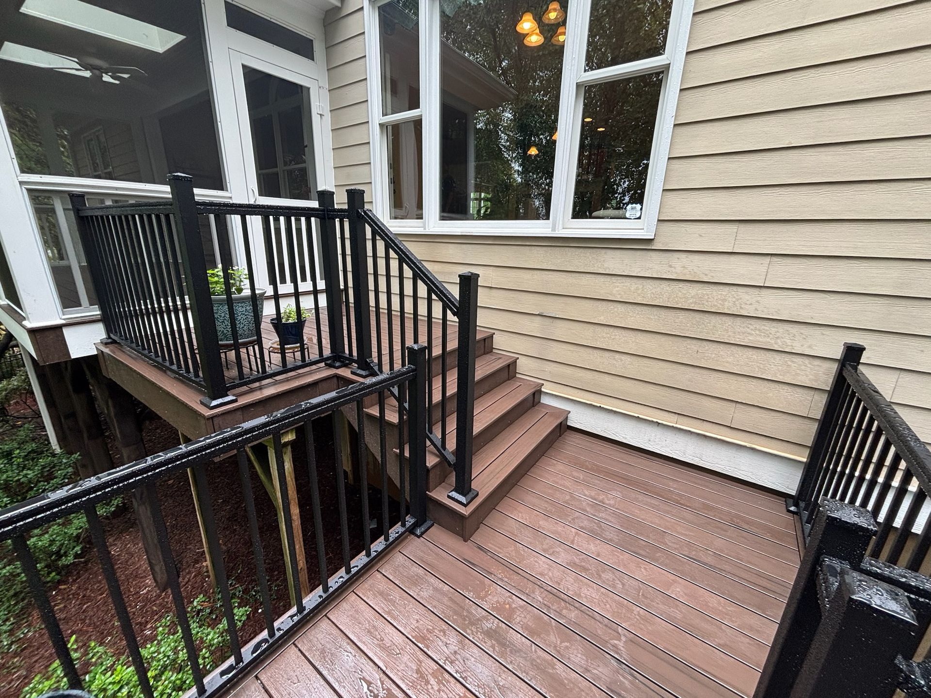 Wooden deck and stairs beside a beige house, with black railings and windows overlooking a landscaped yard