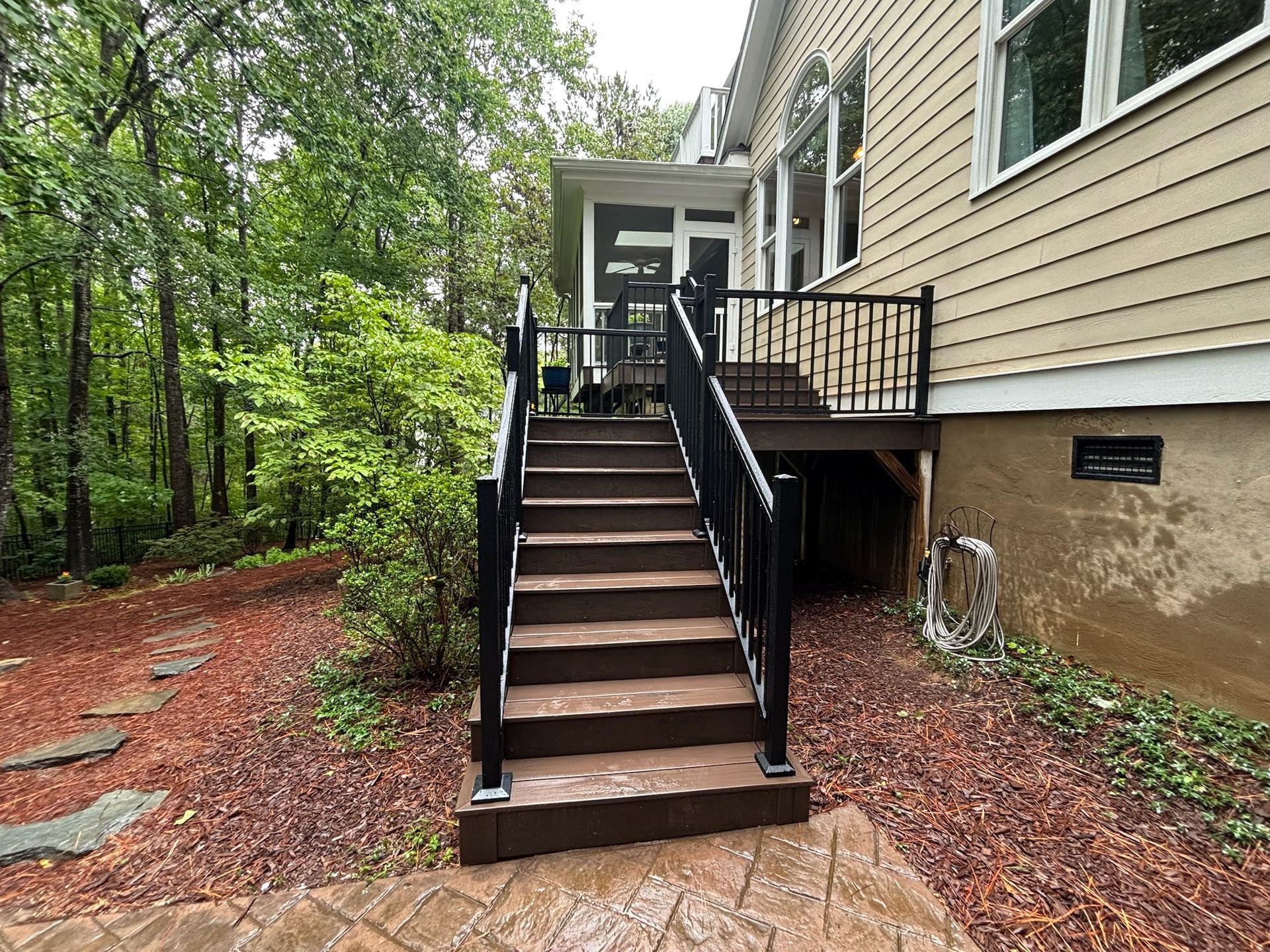 Wooden staircase with black railings beside a beige house, leading up from a wooded yard.