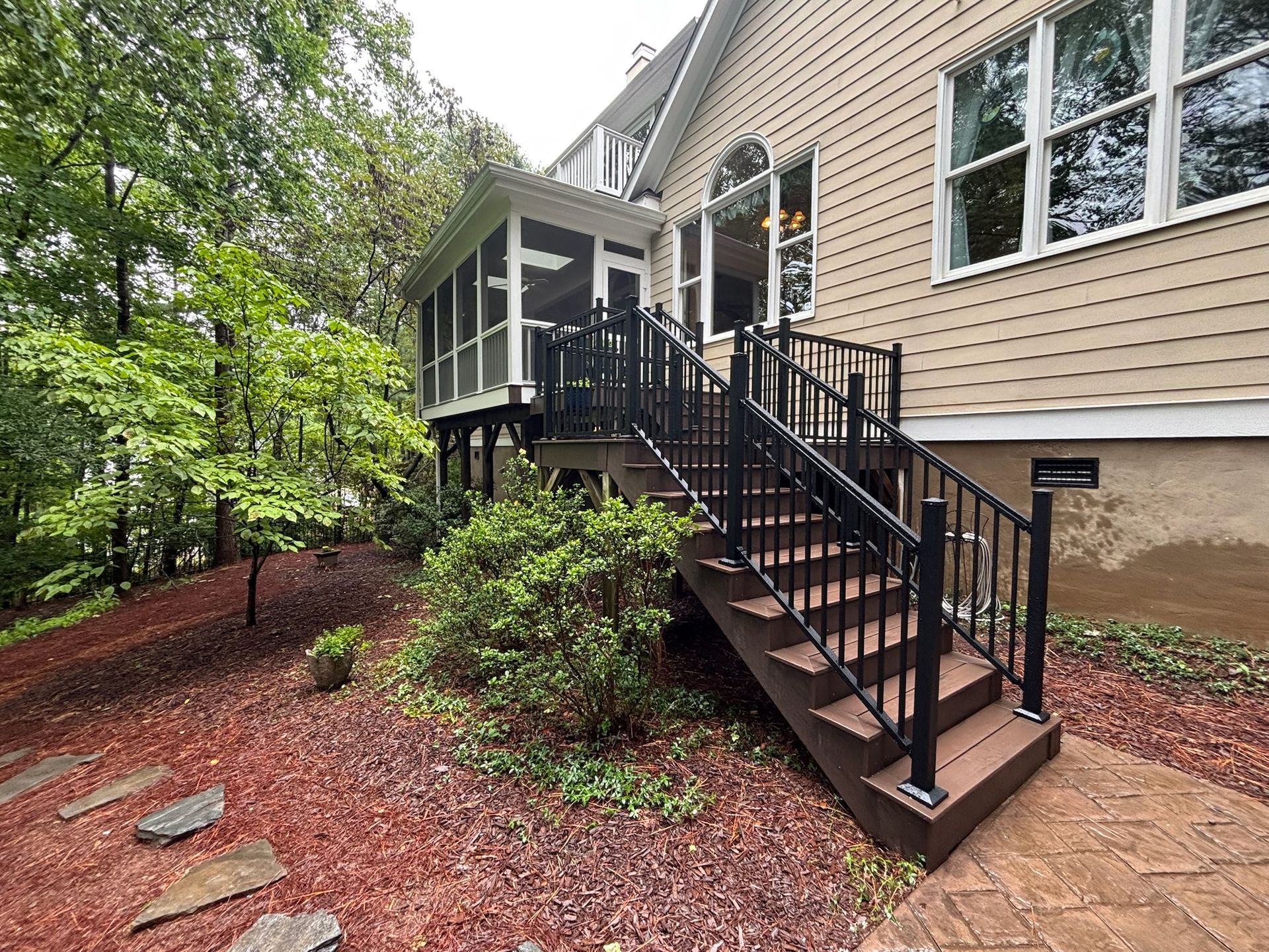 Front entry stairs with black railings leading to a beige house with a screened porch amid trees and mulch beds
